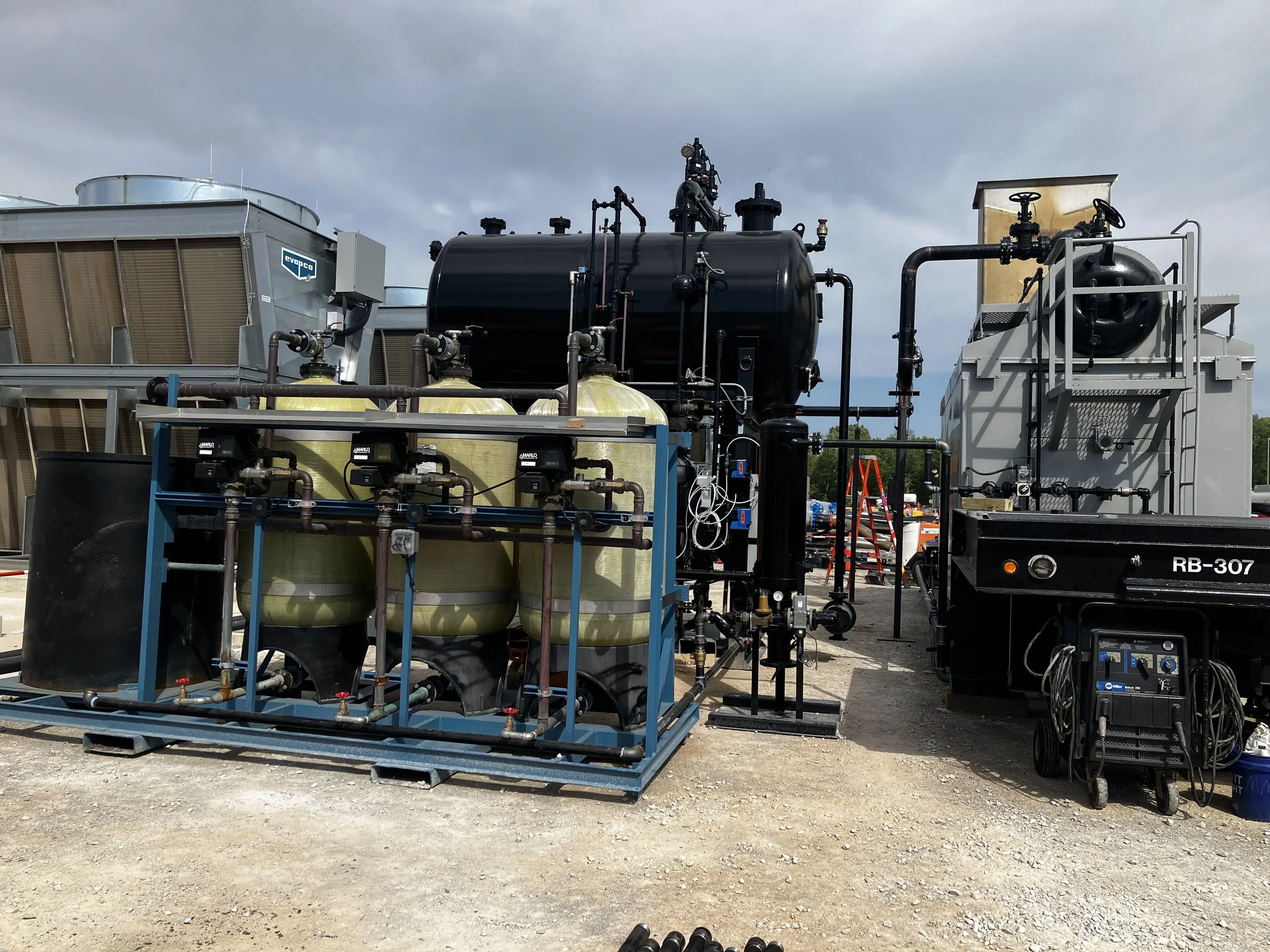 Industrial equipment setup with tanks, pipes, and machinery outdoors on a gravel surface, under cloudy sky.