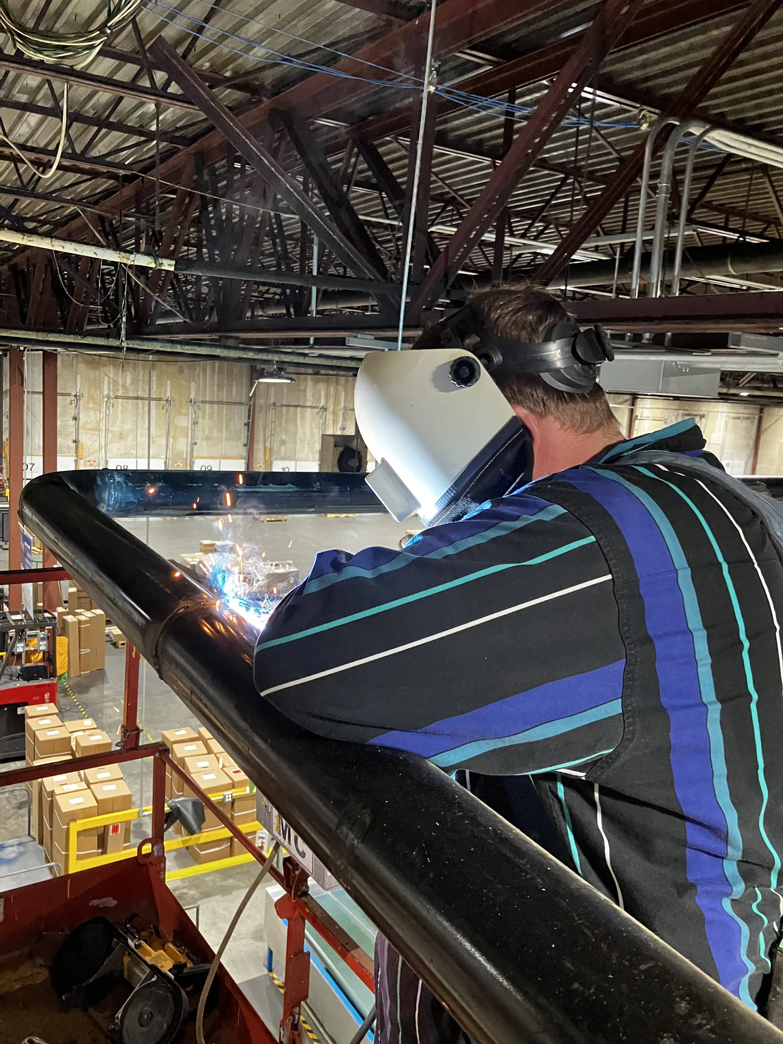 A person welding on a black metal rail inside a warehouse, wearing a white welding helmet and protective gear.