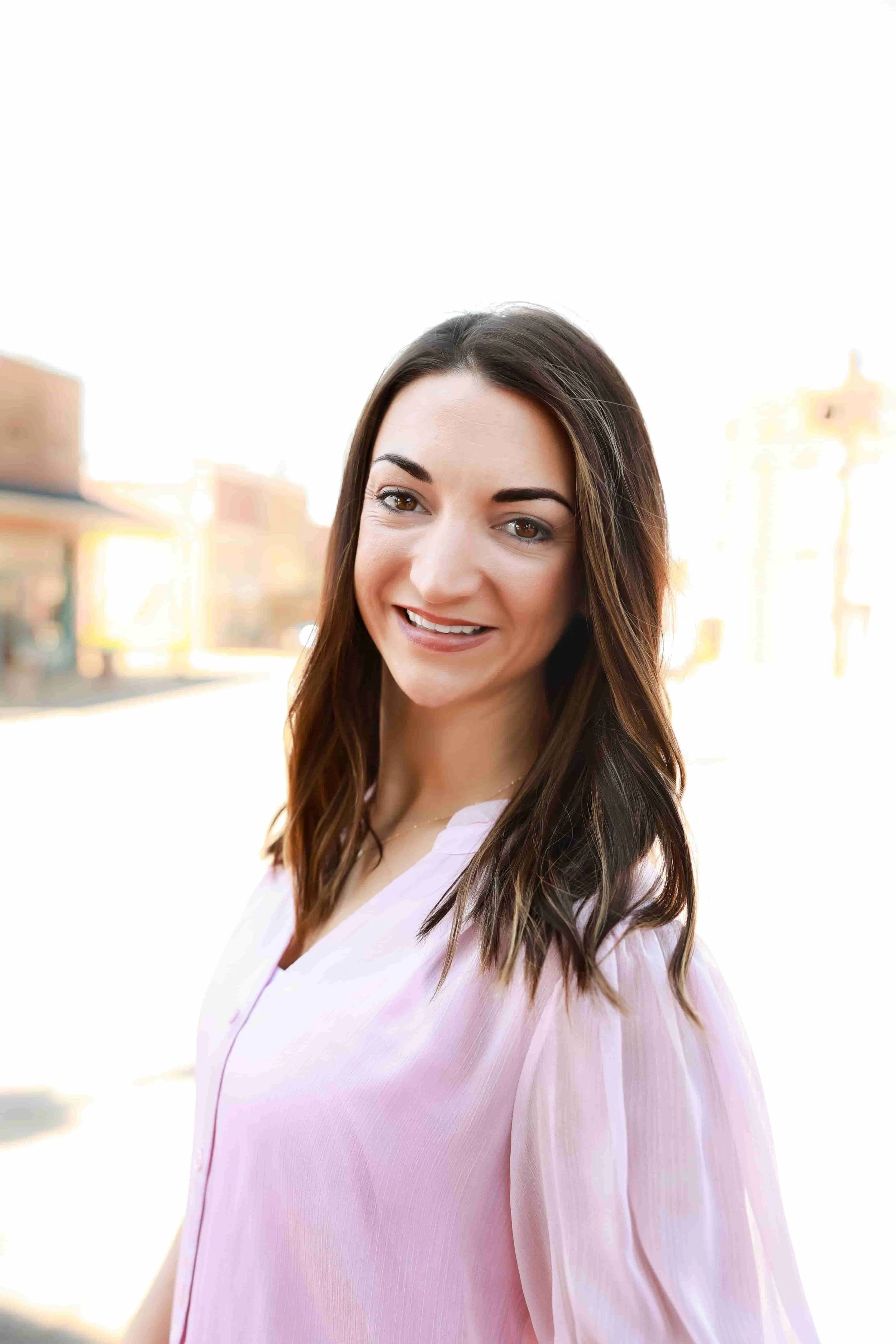 Headshot of Cassi Work - A young woman with long brown hair models outdoors during daylight, wearing a light pink blouse, with a blurred cityscape background.