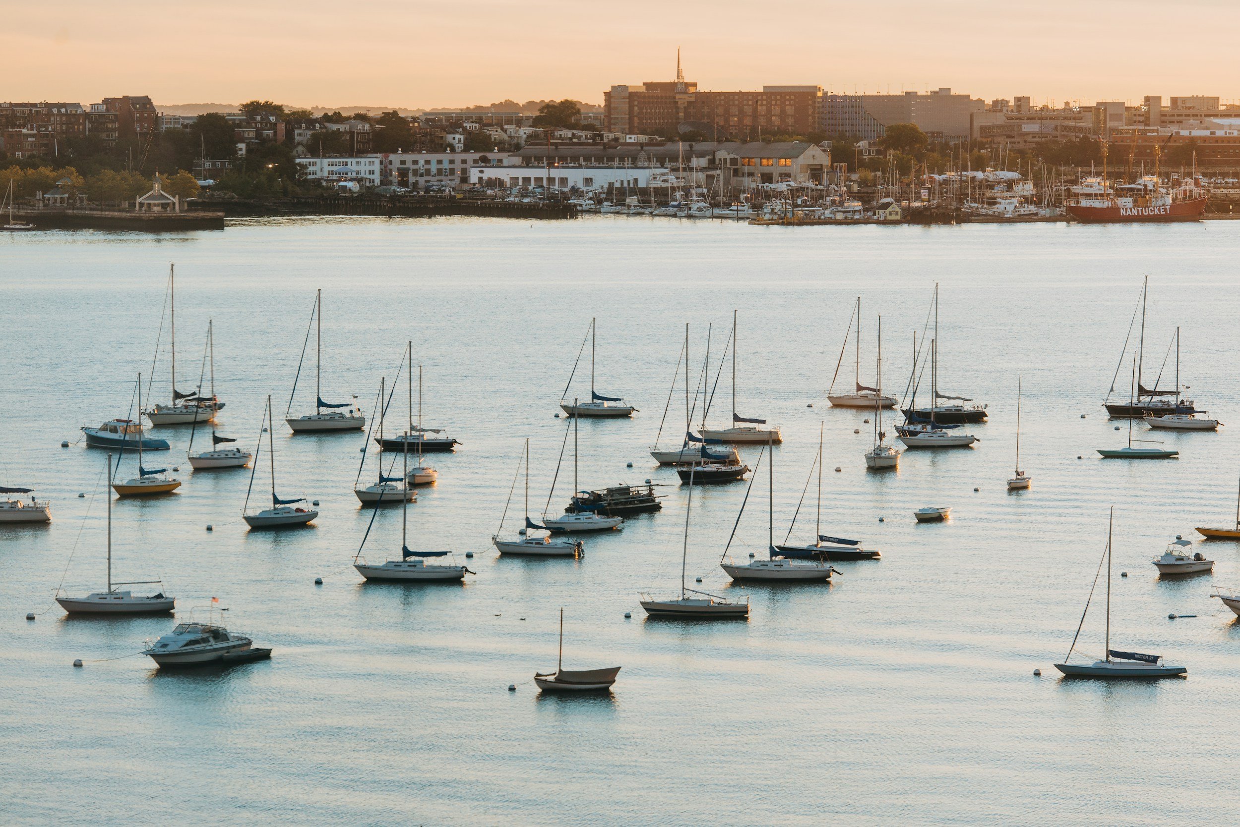 Sailboats anchored in a harbor during sunset with city buildings in the background.