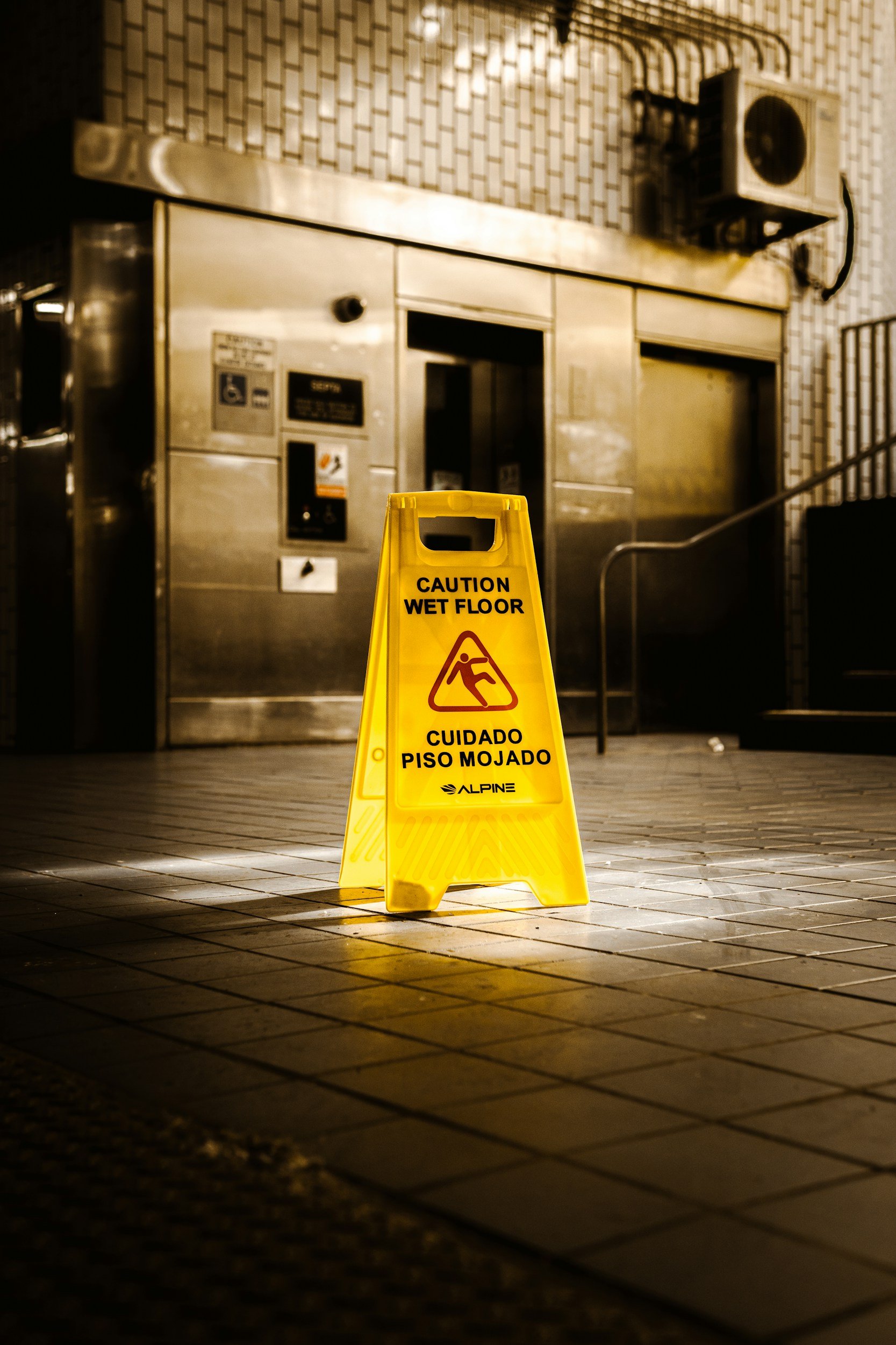 Yellow caution sign on wet floor in front of elevator, with black text in English and Spanish, in an indoor setting with tiled floor and textured wall.