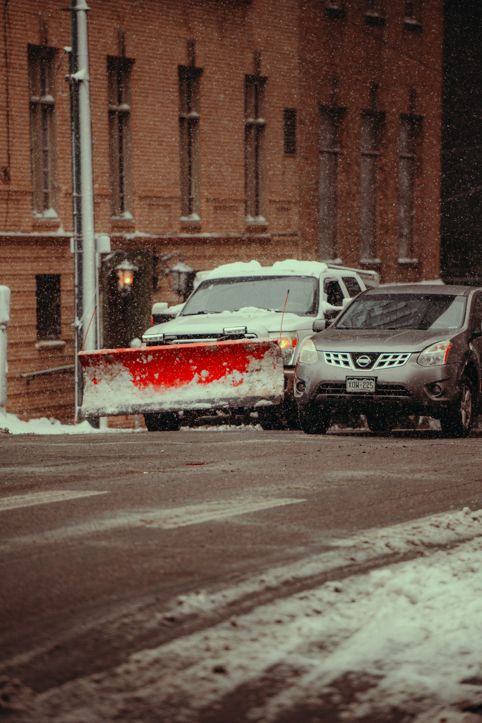 A street scene with snow-covered vehicles and a red snowplow blade clearing the road during snowfall.