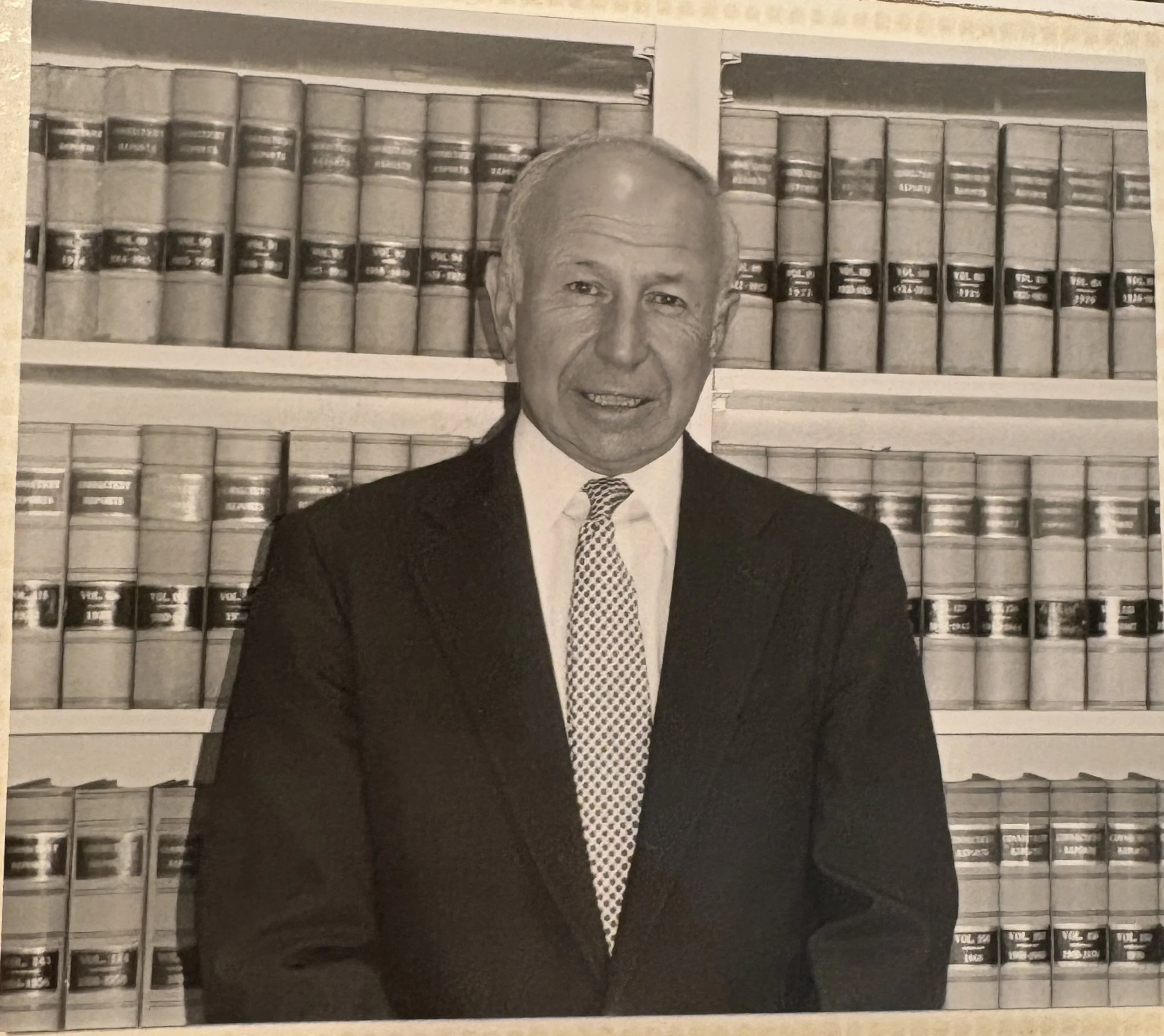 A black and white photo of a middle-aged man in a suit and patterned tie, standing in front of a bookshelf filled with law books.
