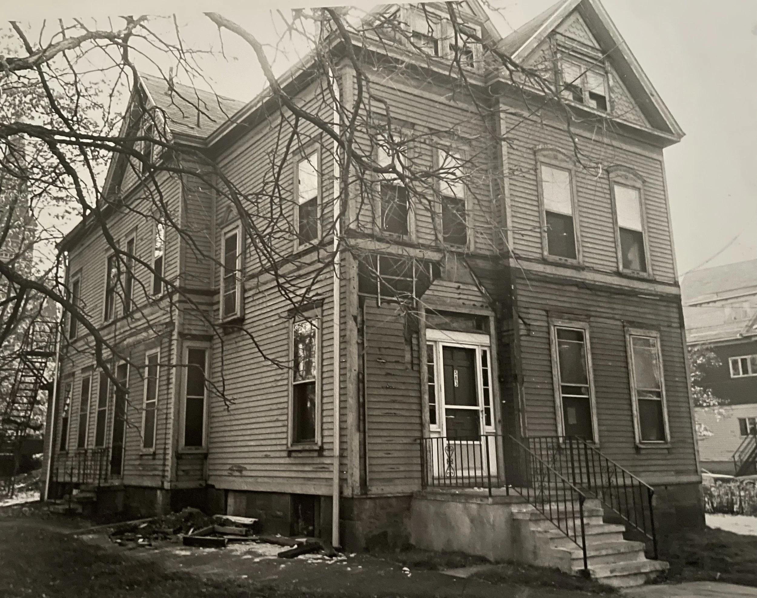 An old, abandoned, two-story wooden house with broken windows, deteriorating siding, and a damaged front porch and stairs. The house is surrounded by leafless trees.