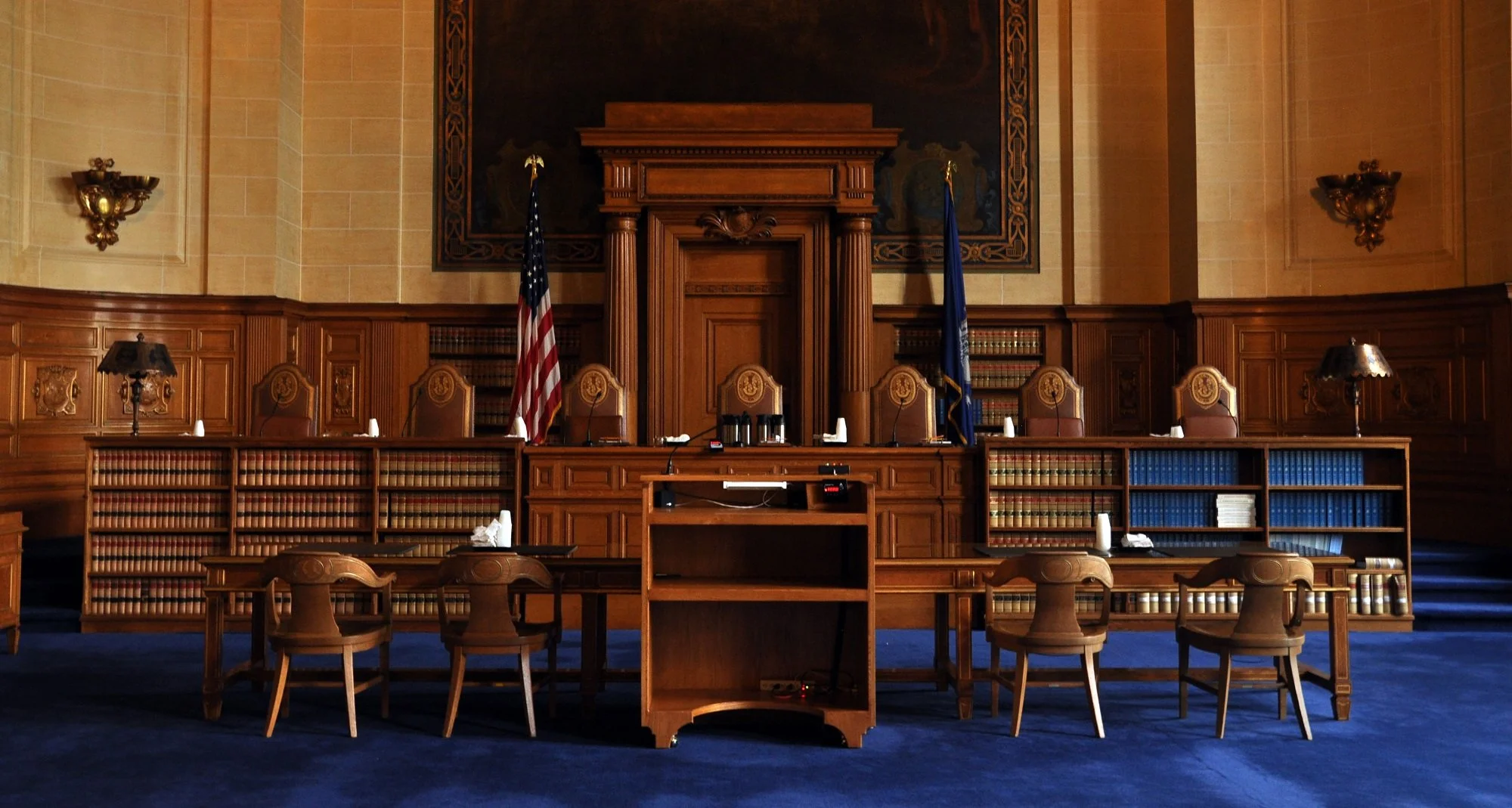 Judicial courtroom with a raised judge's bench, wooden paneling, American flag, blue carpet, and empty chairs.