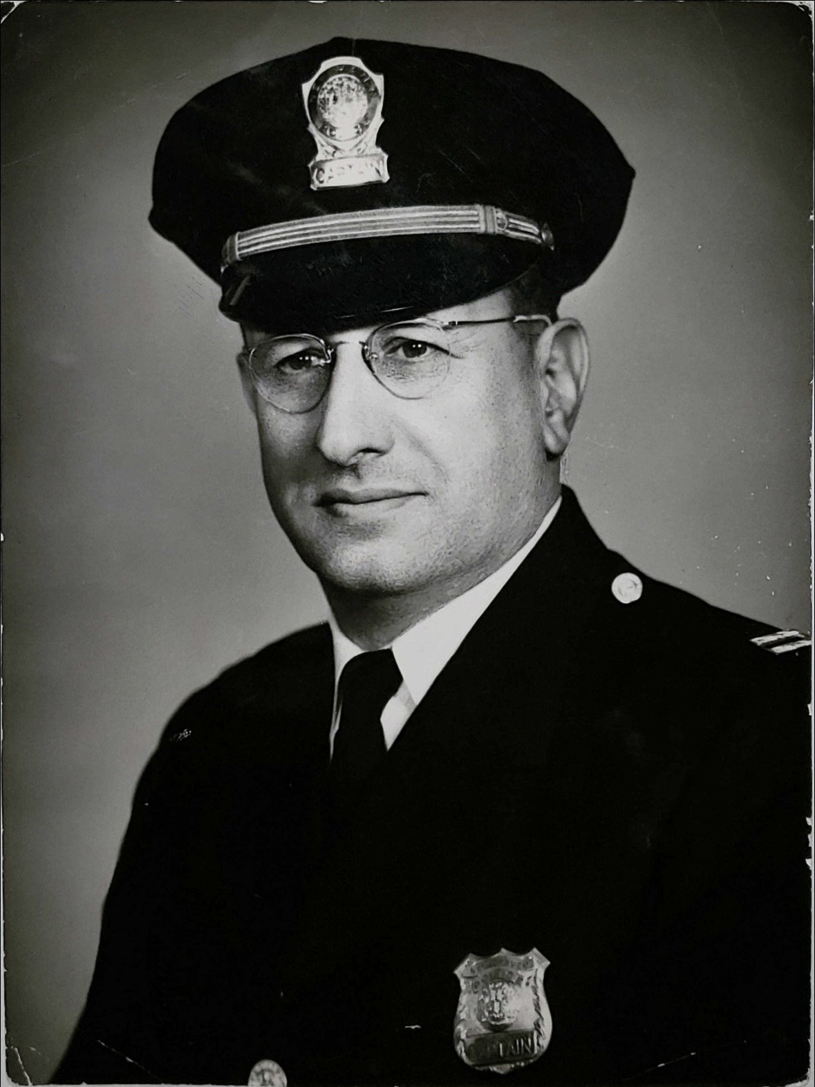 Black and white photograph of a police officer in uniform. The officer is wearing a peaked cap with a badge, glasses, and a police badge on the chest. The background is plain and the officer is looking at the camera with a slight smile.