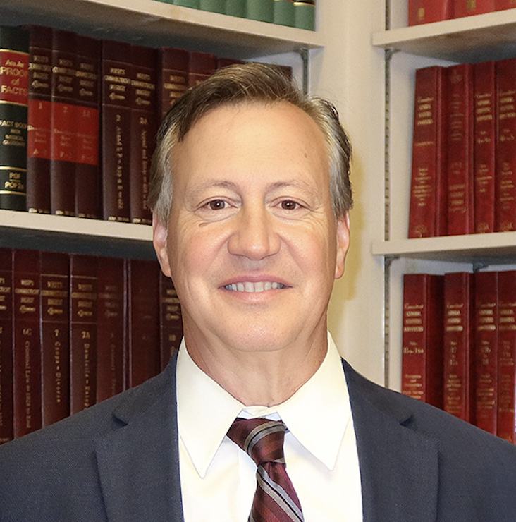 A middle-aged man in a suit and tie smiling, standing in front of bookshelves filled with legal books.