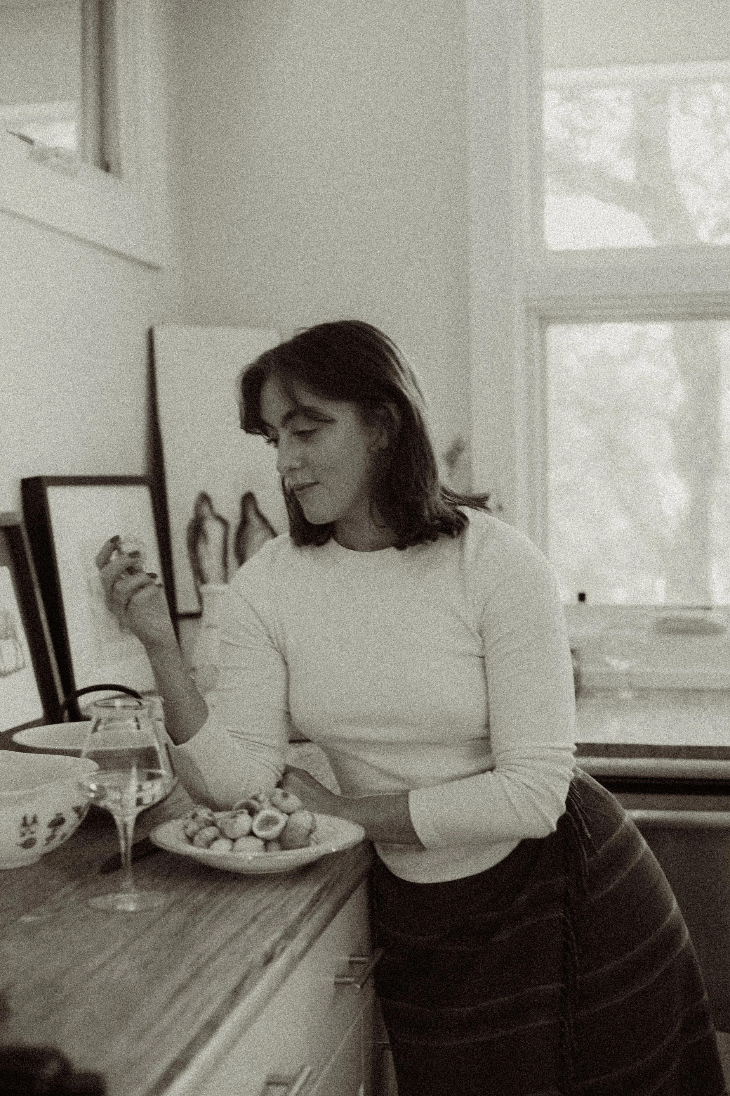 A woman in a white long-sleeve shirt leaning on a kitchen counter, holding a spoon, with a bowl of snacks and a glass of wine nearby. She is holding fresh figs and cooking in the kitchen