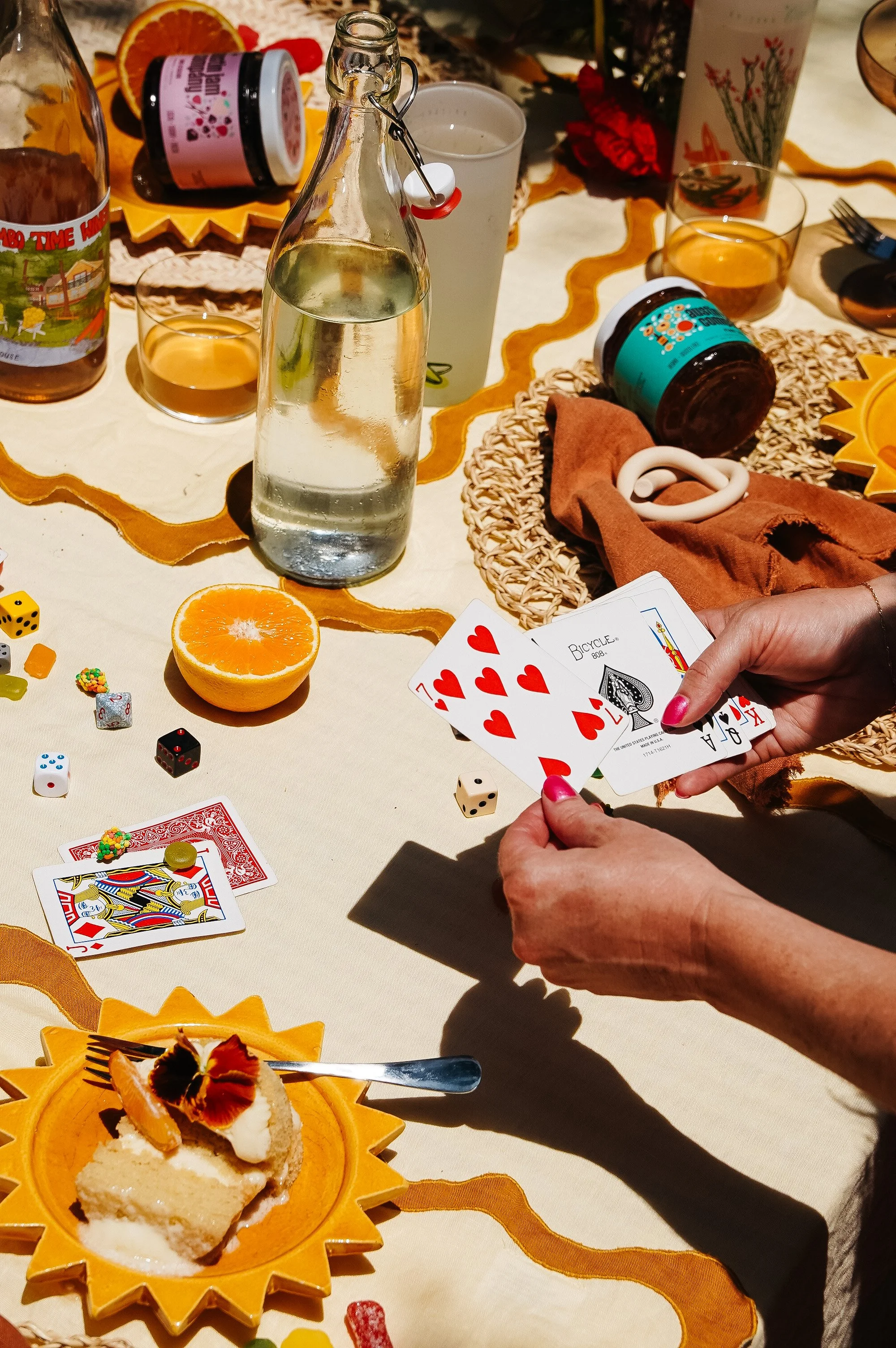 A person holding a hand of playing cards, including the 7, 8, 9, 10, and Jack of hearts, at a table set for a gathering. The table features a half-cut orange, a water bottle, drinks, a dessert, and various game pieces and snacks.