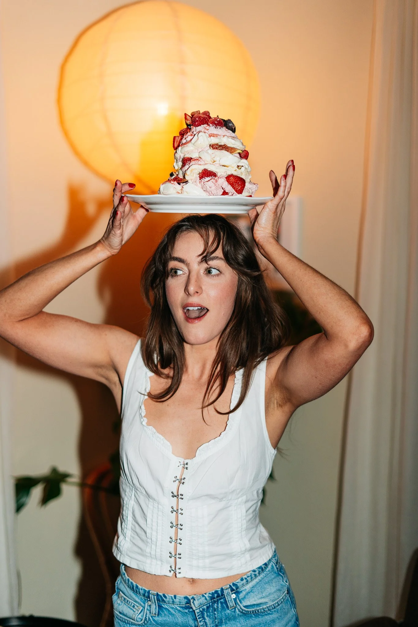 A woman holding a plate of strawberry shortcake above her head with a large lamp in the background.