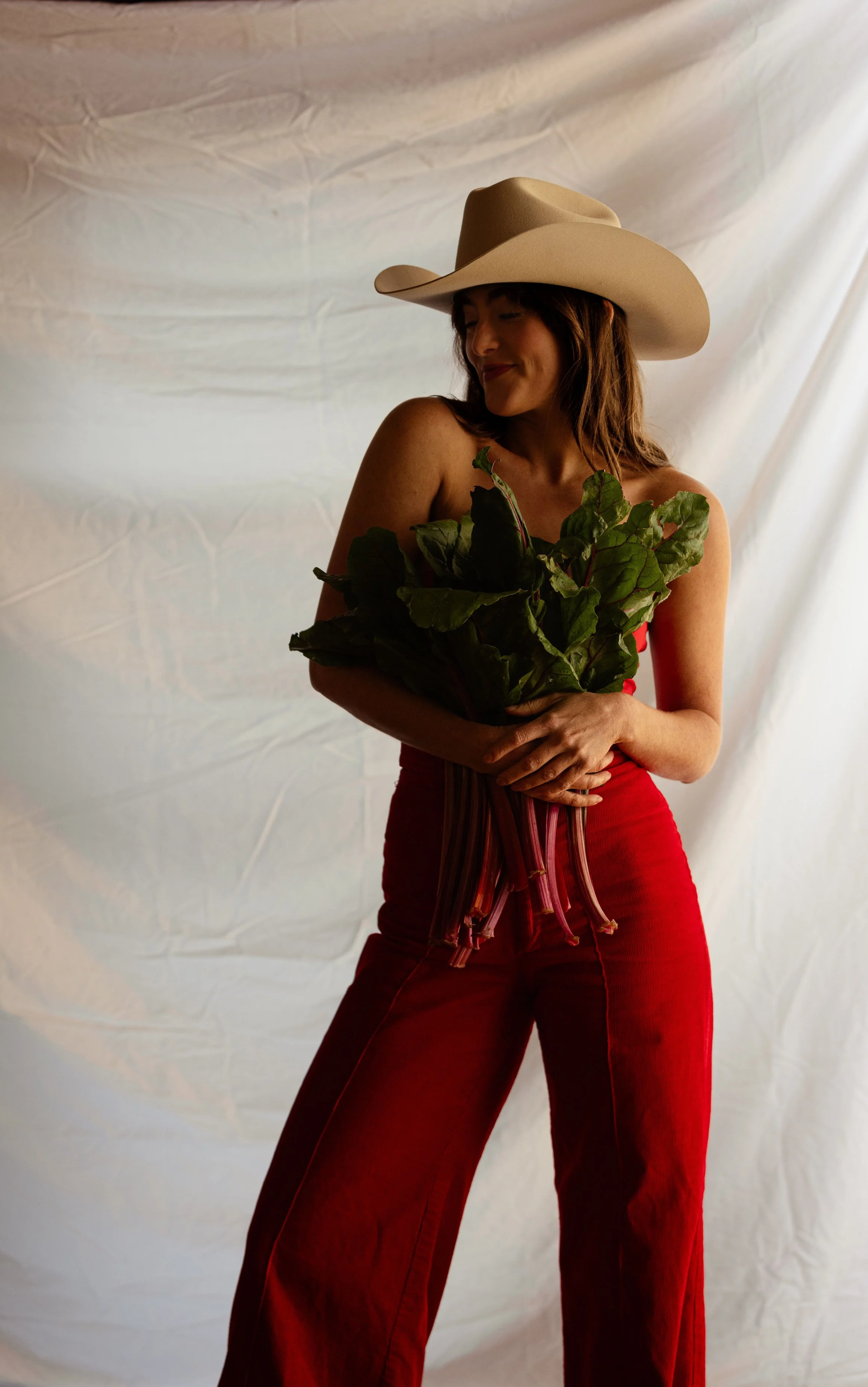 Woman wearing a beige cowboy hat and red outfit holding a bunch of green leafy vegetables against a plain, light-colored background.