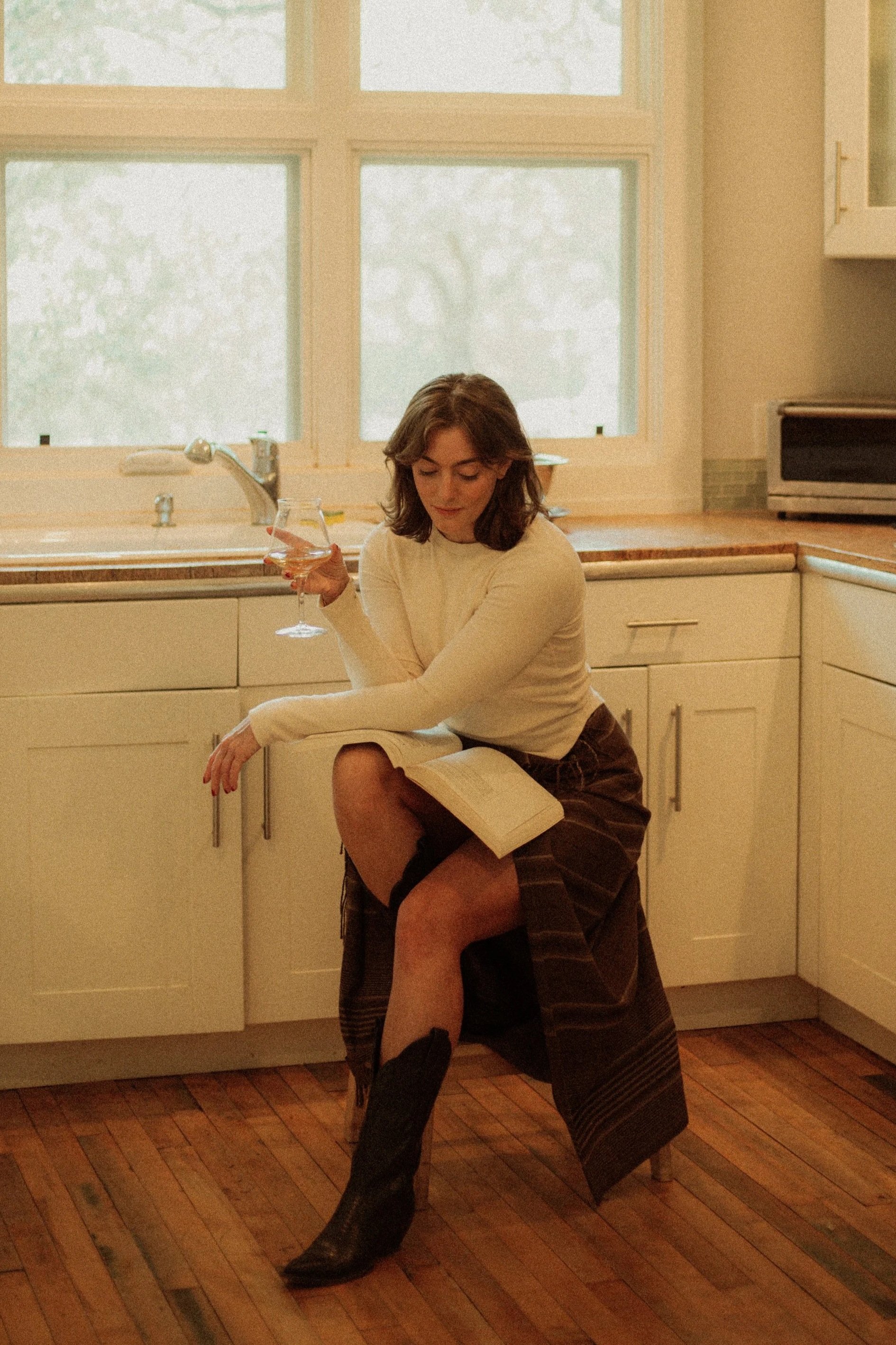 A woman sitting on a kitchen stool, wearing a cowboy boots, holding a glass and a book, in a well-lit kitchen with large windows.