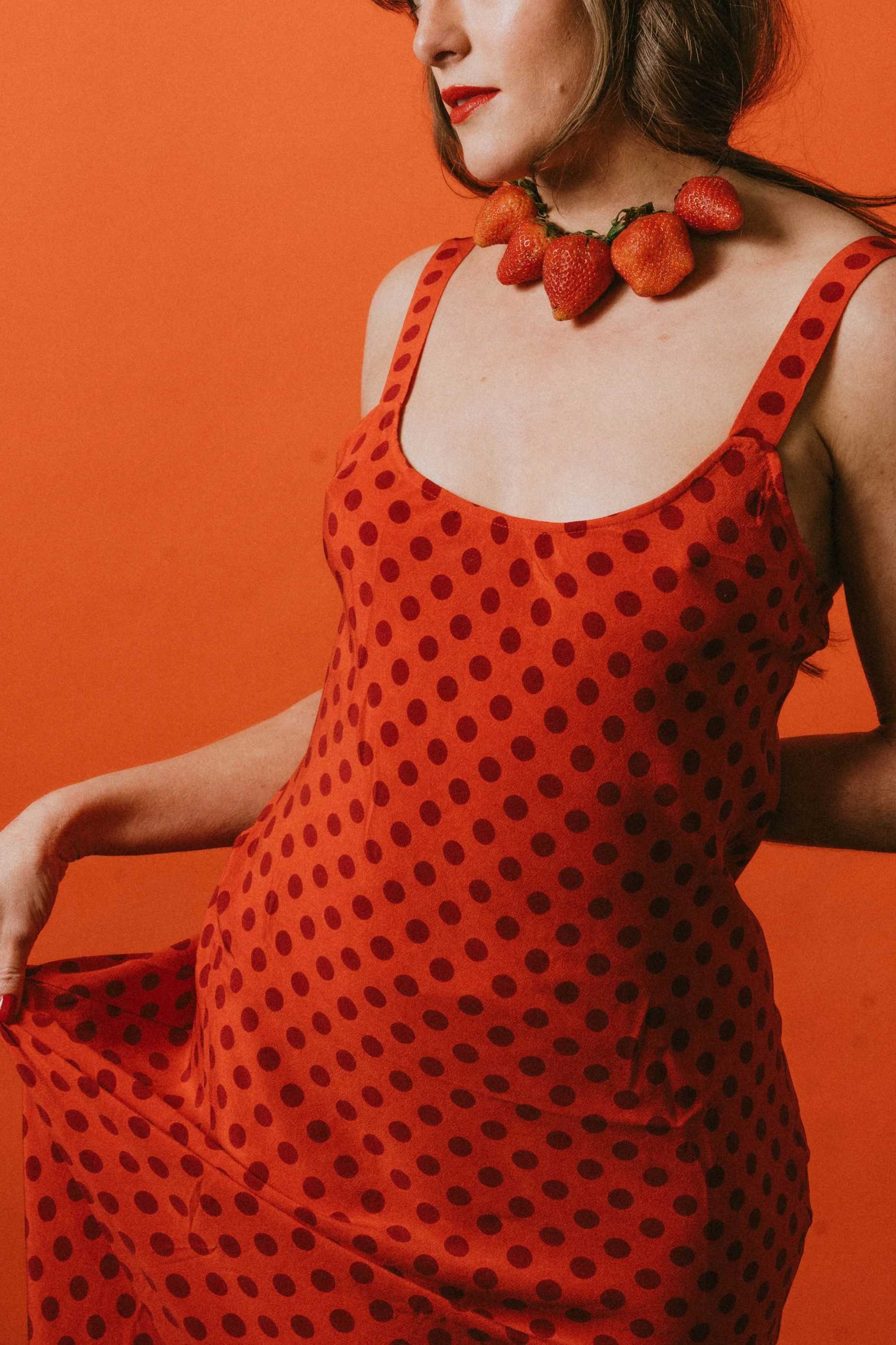 A woman wearing a red polka dot dress and strawberry necklace stands against an orange background. Creative Food Styling Photoshoot