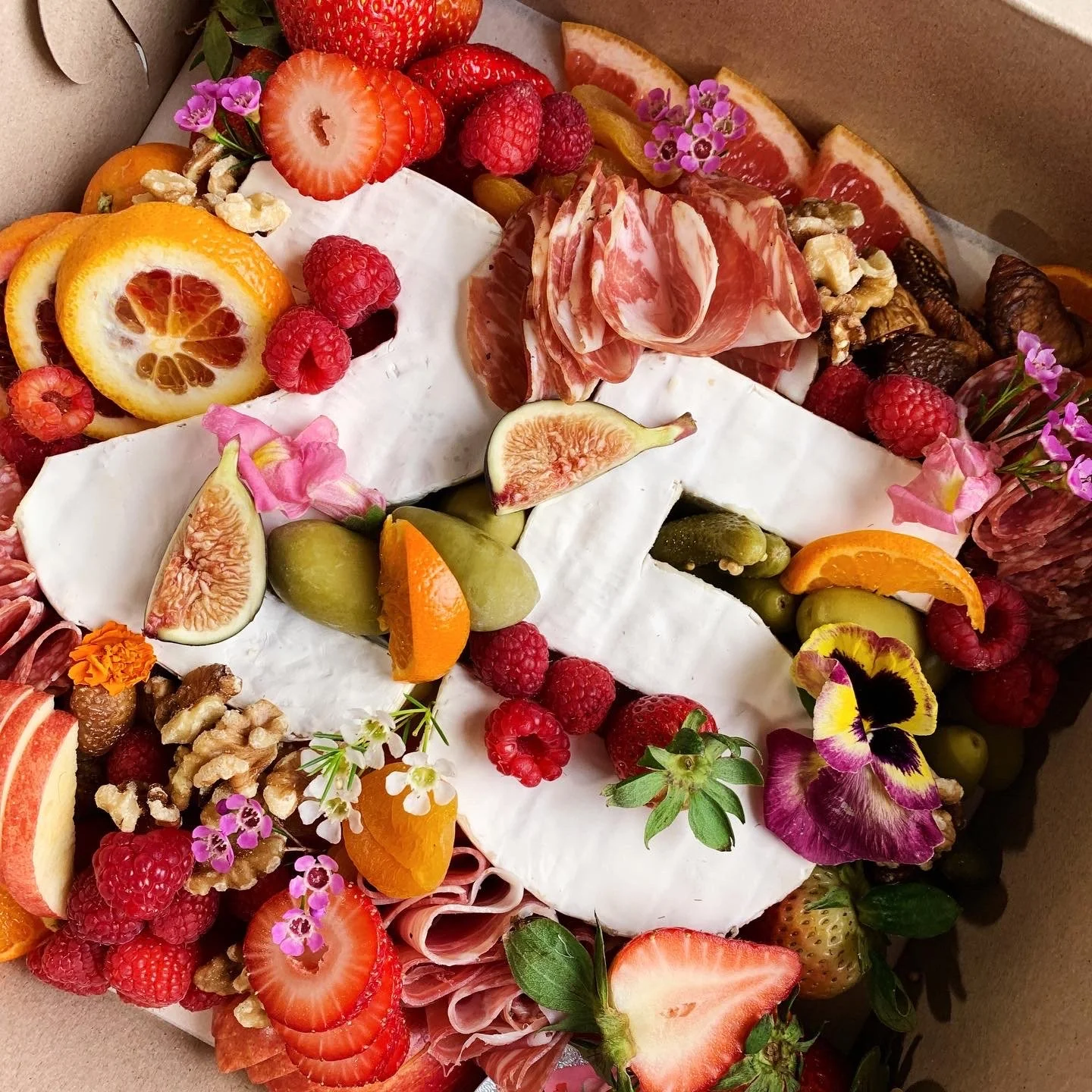 A cheese and charcuterie platter decorated with fresh fruits like strawberries, raspberries, figs, oranges, and grapes, as well as nuts and edible flowers.