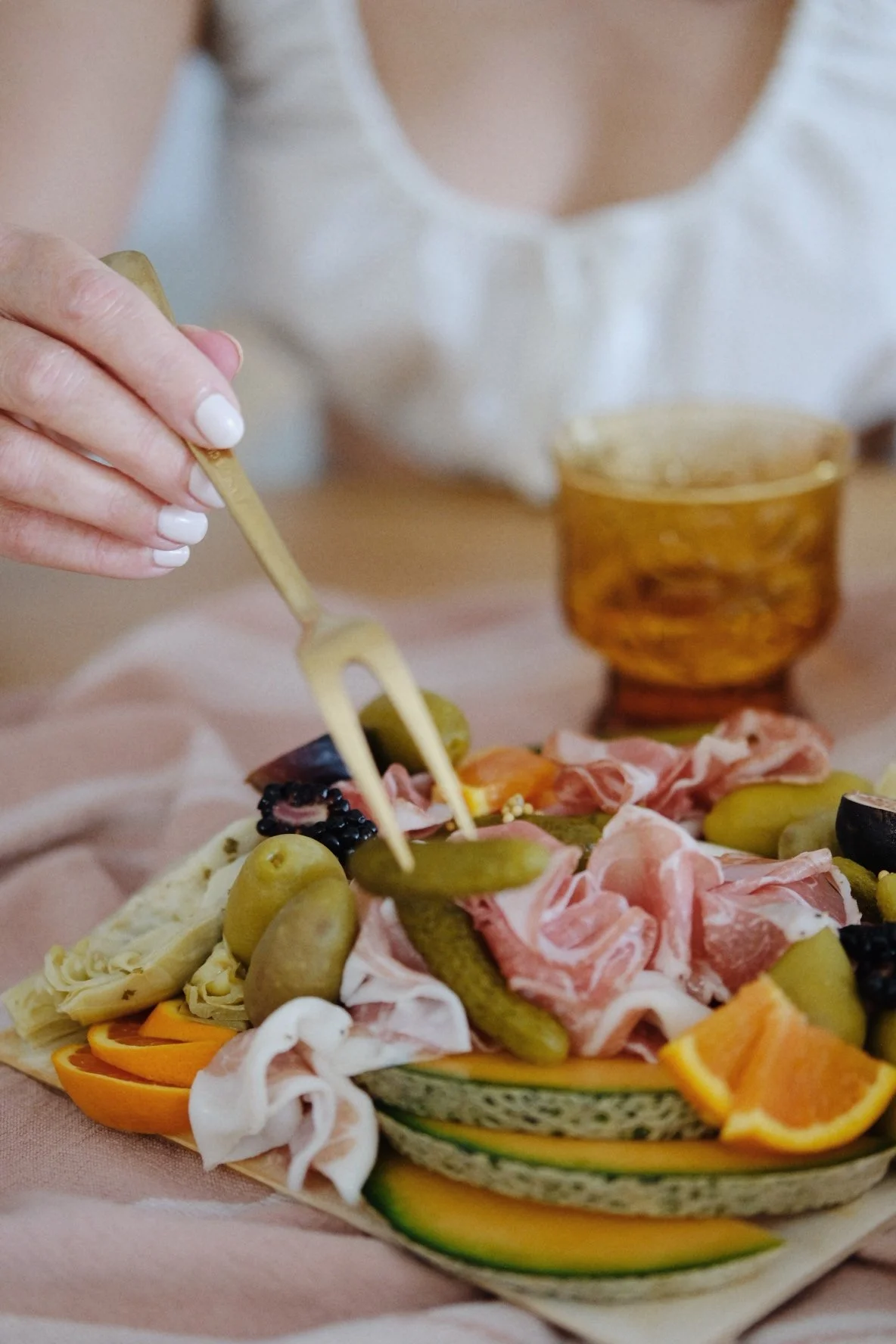 A person with white painted nails is using a small fork to pick up a green olive from a charcuterie board. The board contains various cheeses, sliced meats, grapes, blackberries, and sliced oranges—resting on a pink cloth with a glass of amber-colore
