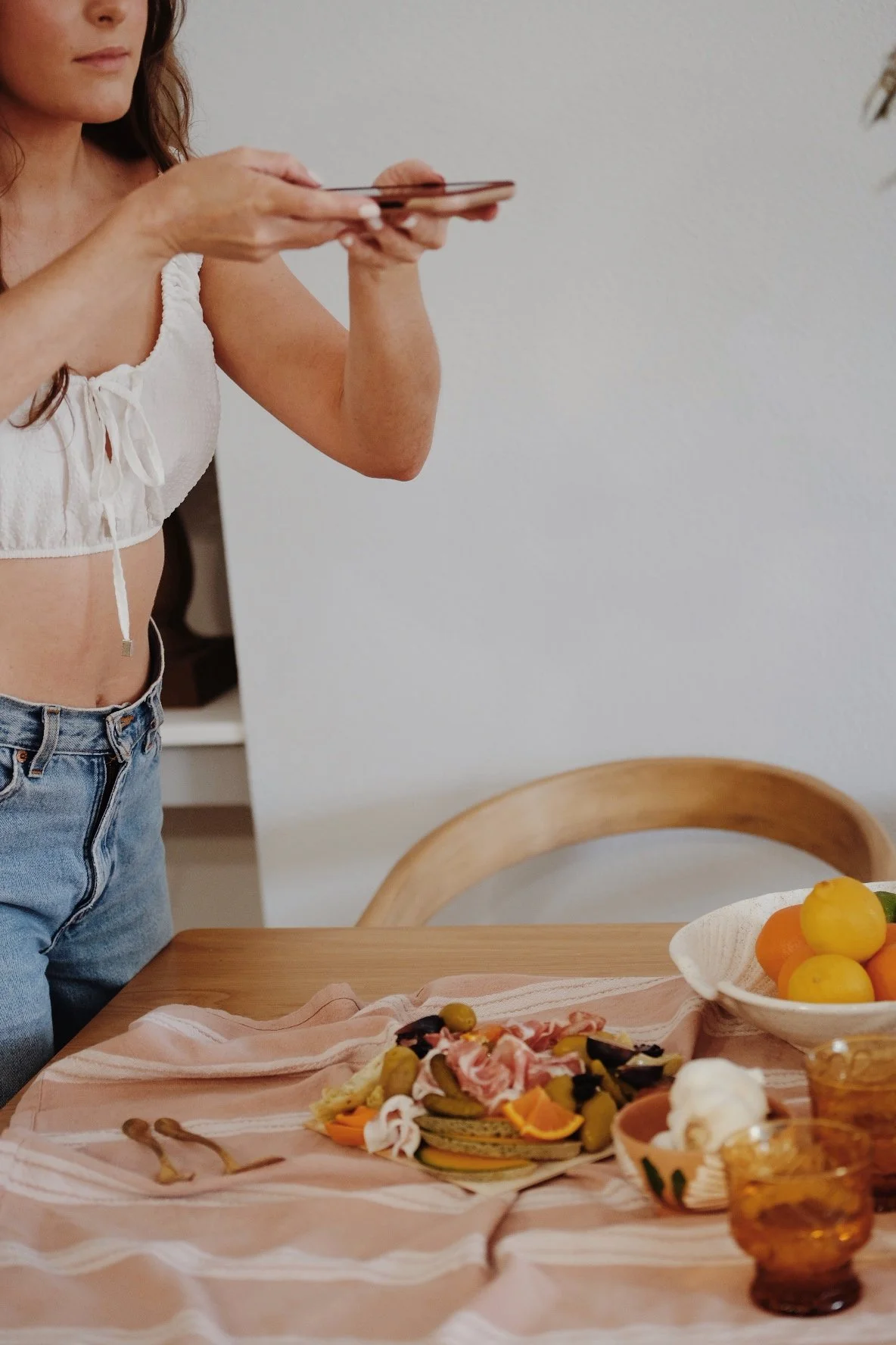 A woman taking a photo of a table with food, including a charcuterie board, a bowl of fruit, and two glasses of amber-colored drink.