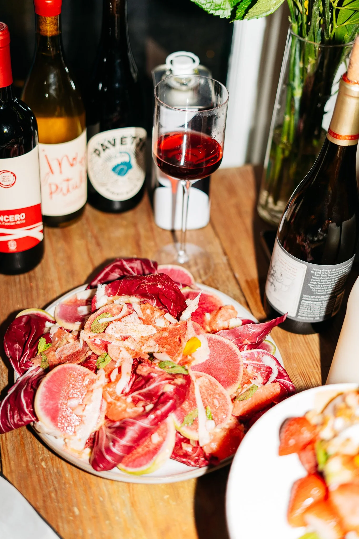 A wooden table with a plate of sliced citrus fruits topped with herbs, a glass of red wine, and various bottles of wine and alcohol in the background, along with a vase of green leaves.
