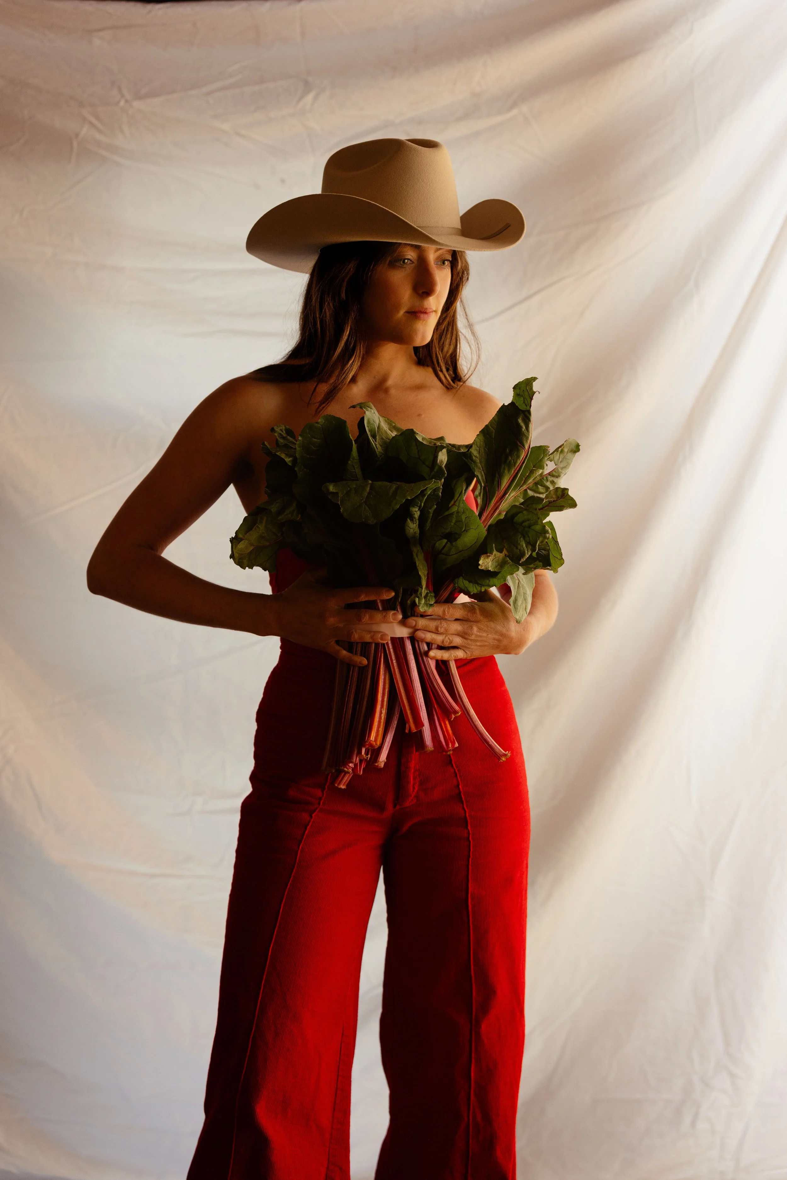 A woman in a beige cowboy hat and red pants holding a bunch of leafy greens, standing against a neutral backdrop.