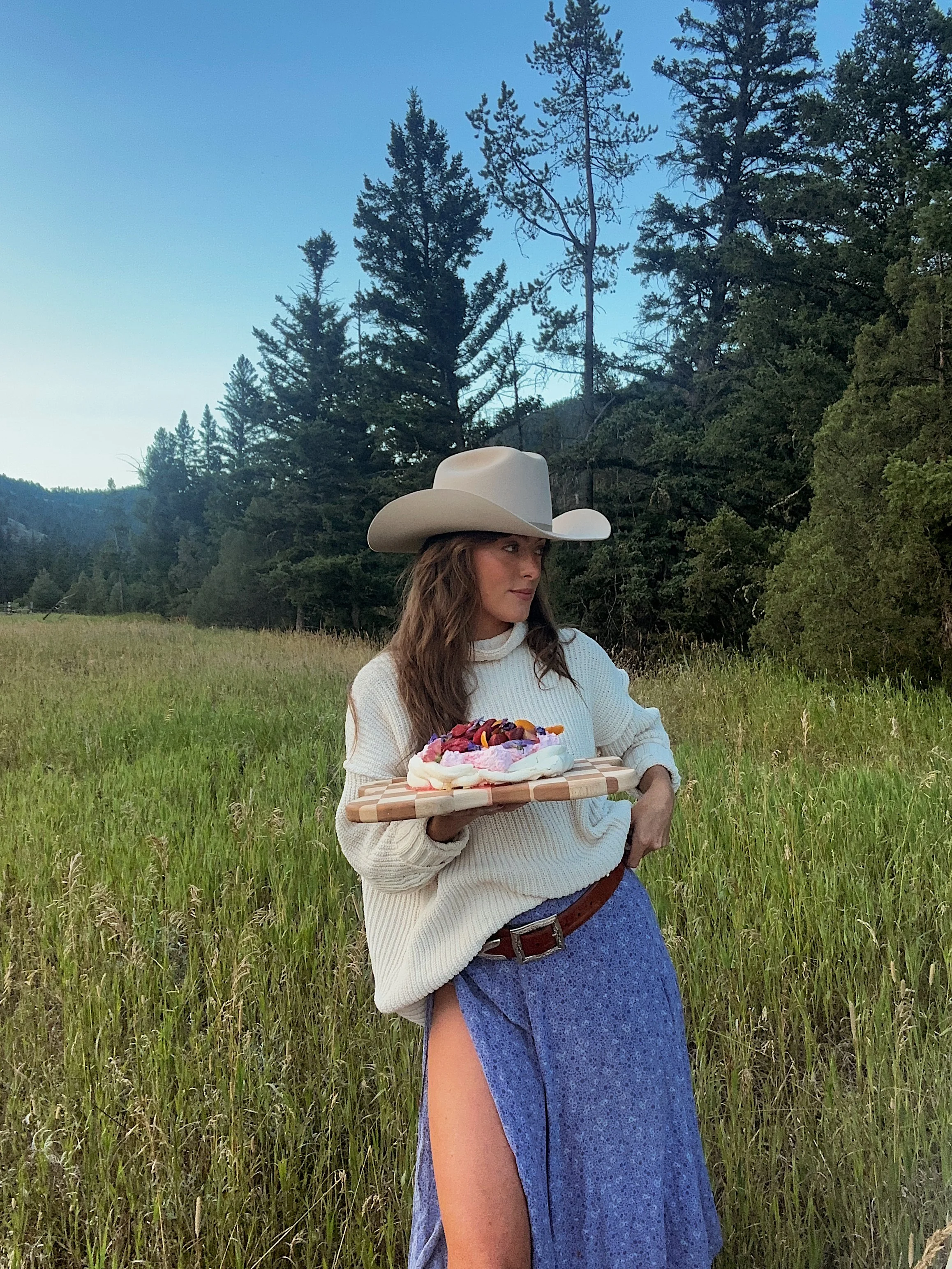 A woman wearing a cowboy hat and white sweater walking in a grassy field and holding a wooden tray with a colorful dessert or cake