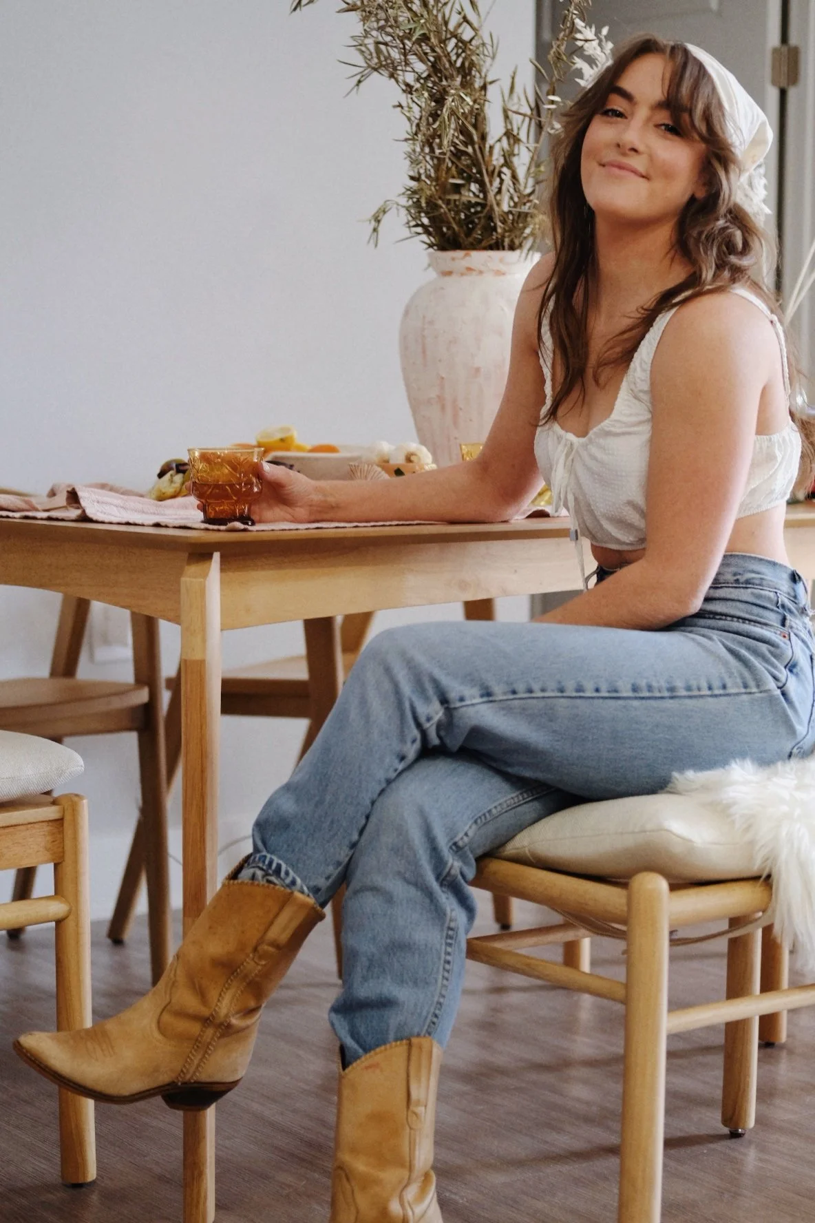 A woman sitting on a wooden chair at a dining table, smiling. She is wearing a white tank top, blue jeans, and tan cowboy boots. The table behind her has a pink cloth, a large pink vase with dried plants, and a plate of fruit.