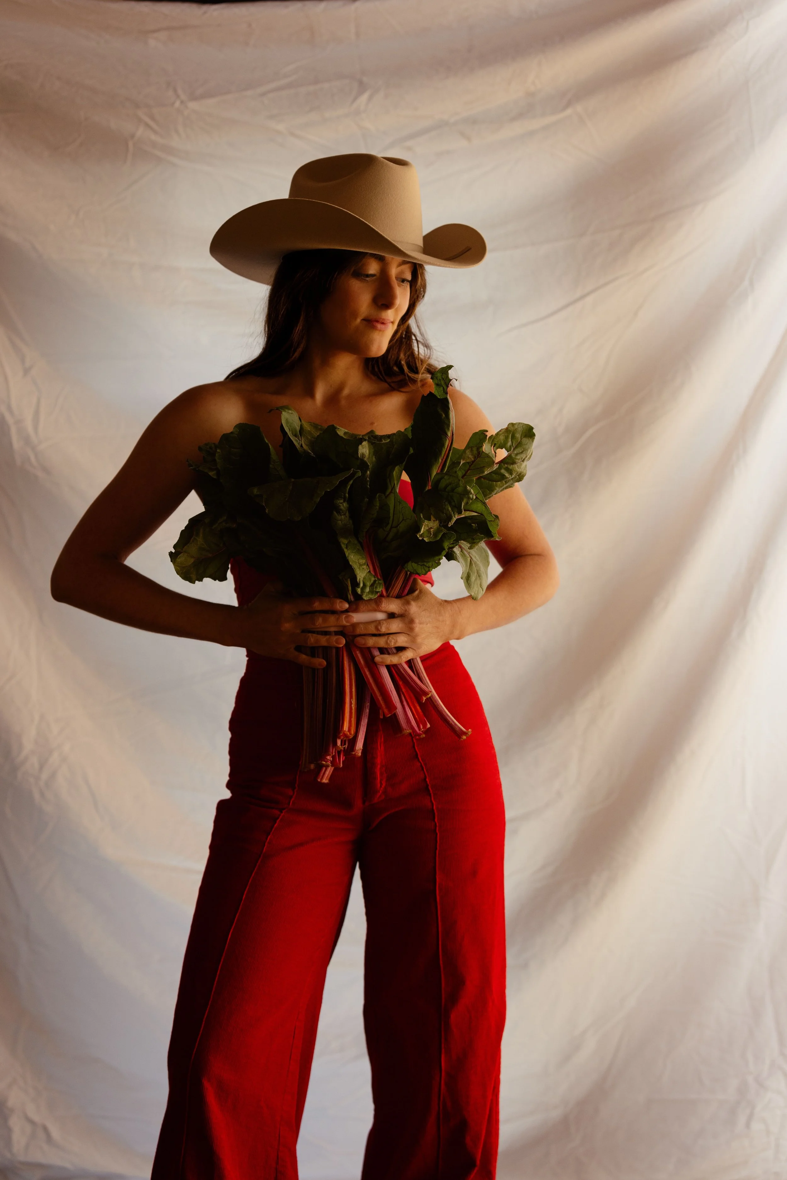 A woman wearing a cowboy hat and red pants, holding a bunch of Swiss chard leaves, standing against a plain backdrop.