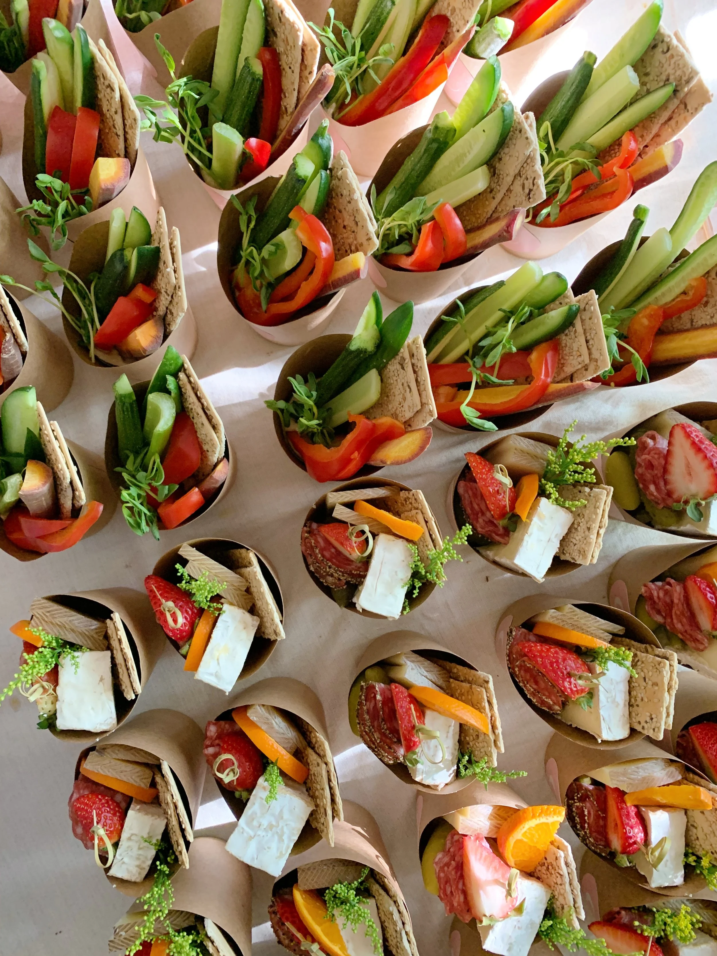 Assorted mini sandwiches and vegetable cups arranged on a table, featuring sliced strawberries, oranges, and fresh vegetables, garnished with herbs.