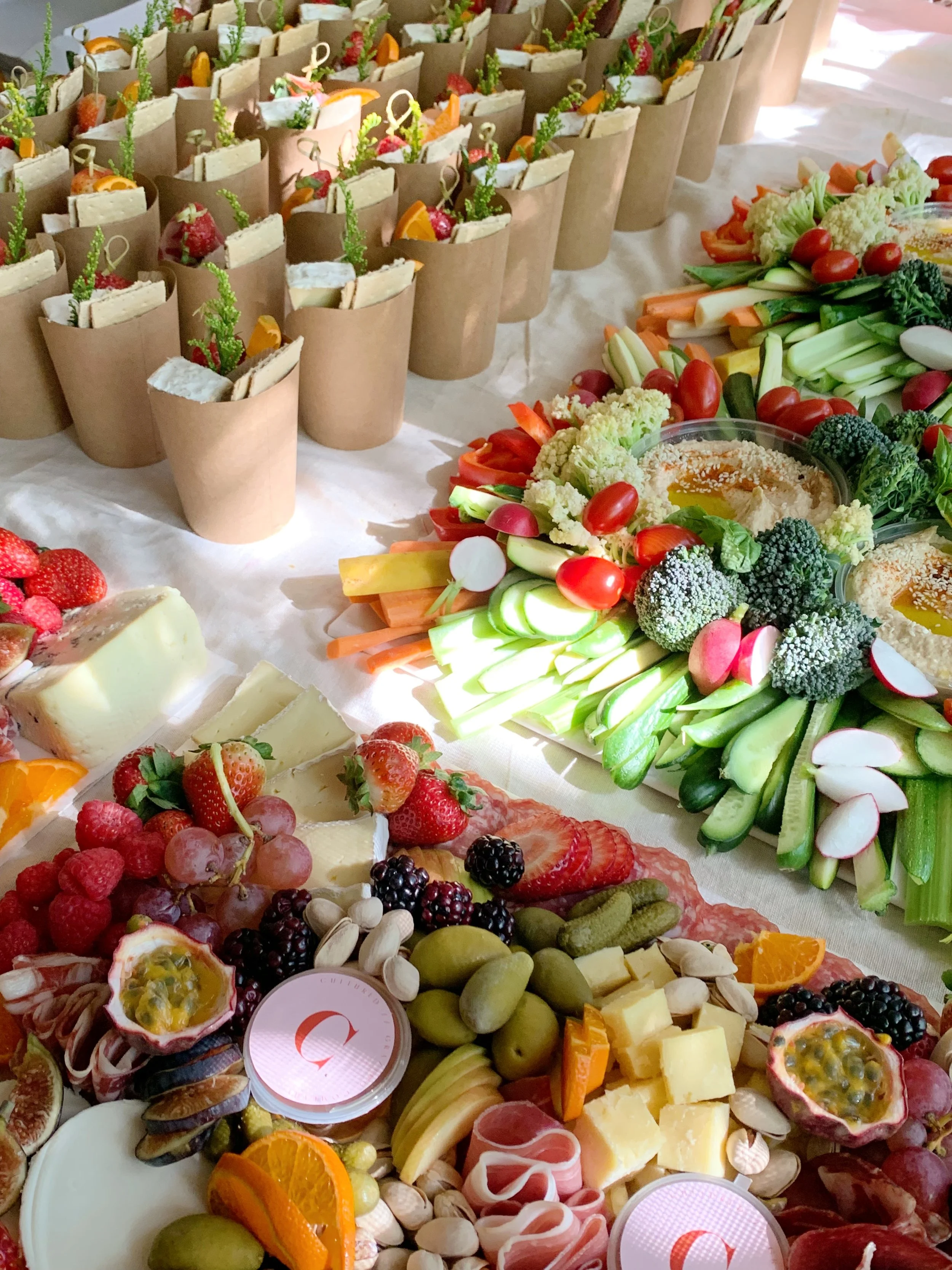 A table with an assortment of finger foods including sandwiches in paper cones, fresh vegetables, a cheese and charcuterie platter with grapes, strawberries, blackberries, pistachios, and passion fruit, and vegetable decorations around hummus.