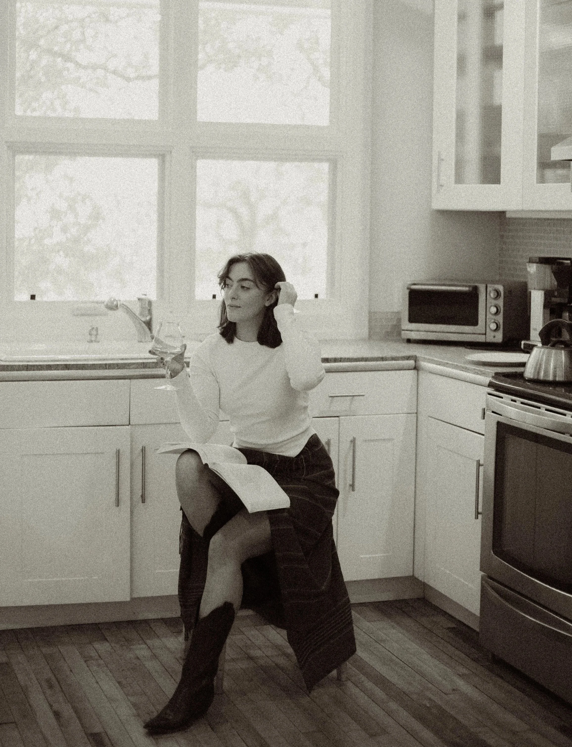 A woman relaxing in a kitchen, holding a glass of wine in one hand and an open cookbook in her lap, near a window with trees outside.