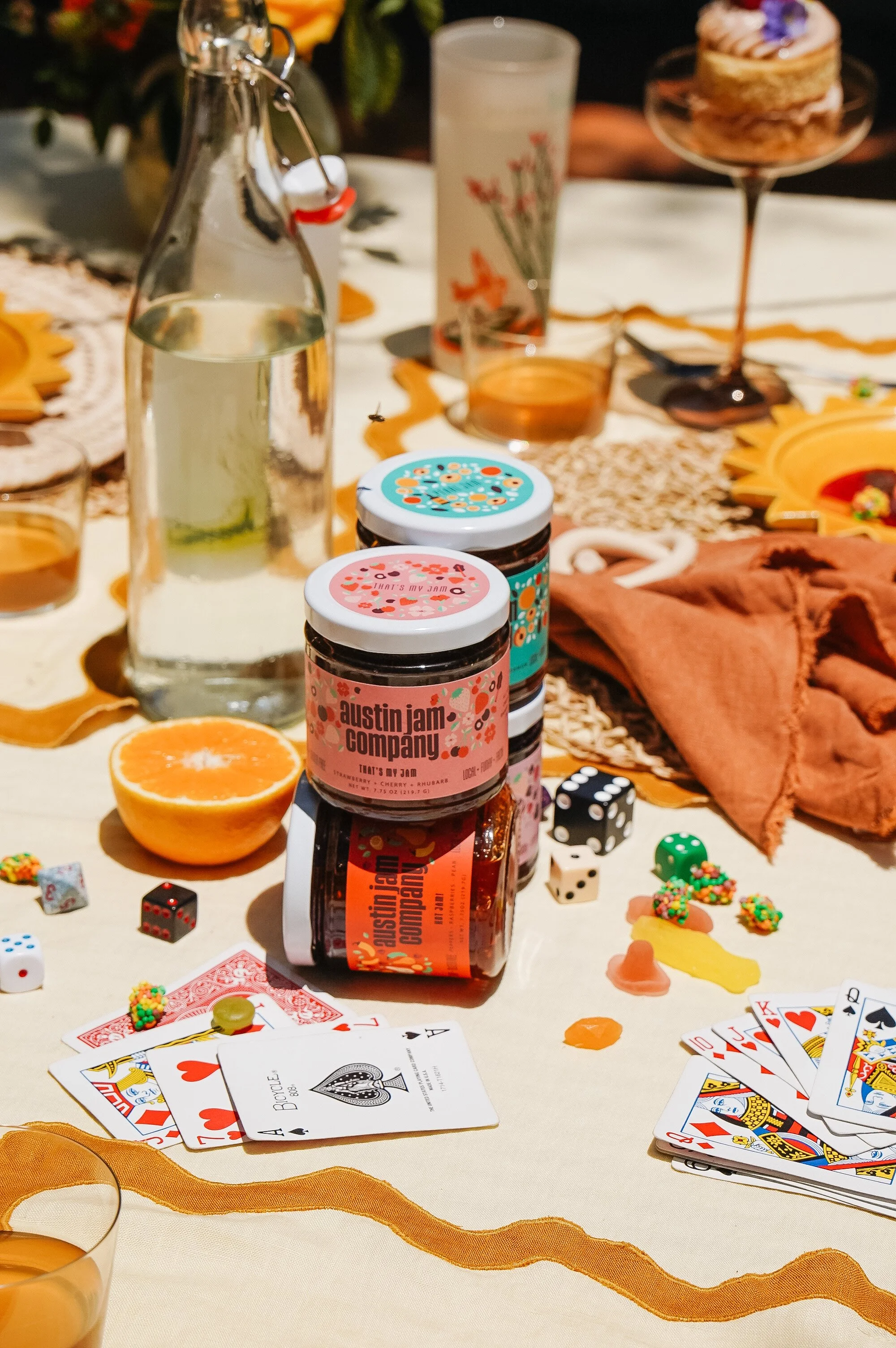 A table with playing cards, dice, jars of Austin Jam Company preserves, a halved orange, a glass bottle of water, and various party items on a beige tablecloth.