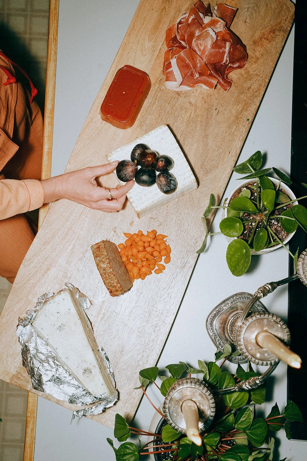 A wooden table with several food items, including a wedge of blue cheese, fig fruits, orange berries, sliced cured meat, a block of honey, and a soap bar. There are also potted plants and decorative glass bottles with straws on the table.