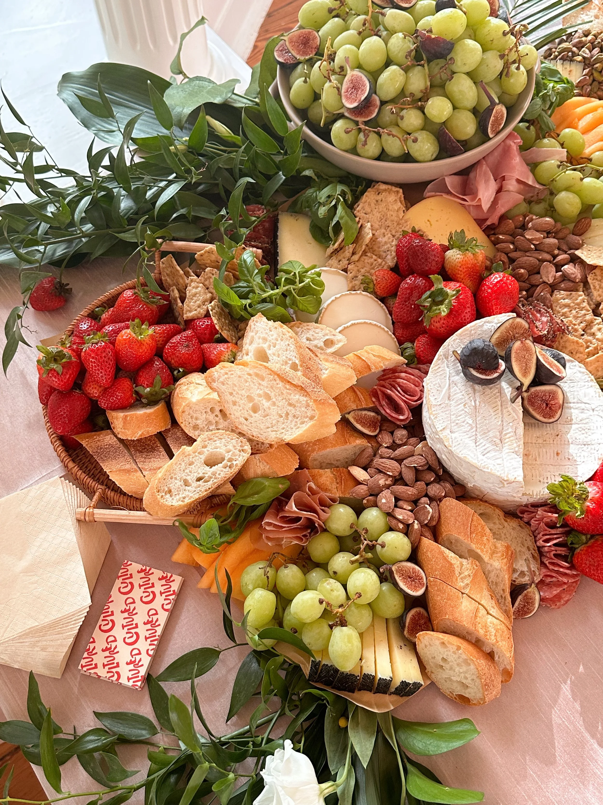 A charcuterie board with grapes, strawberries, cheese, bread, nuts, and meats, surrounded by green foliage and a bowl of green grapes with figs.