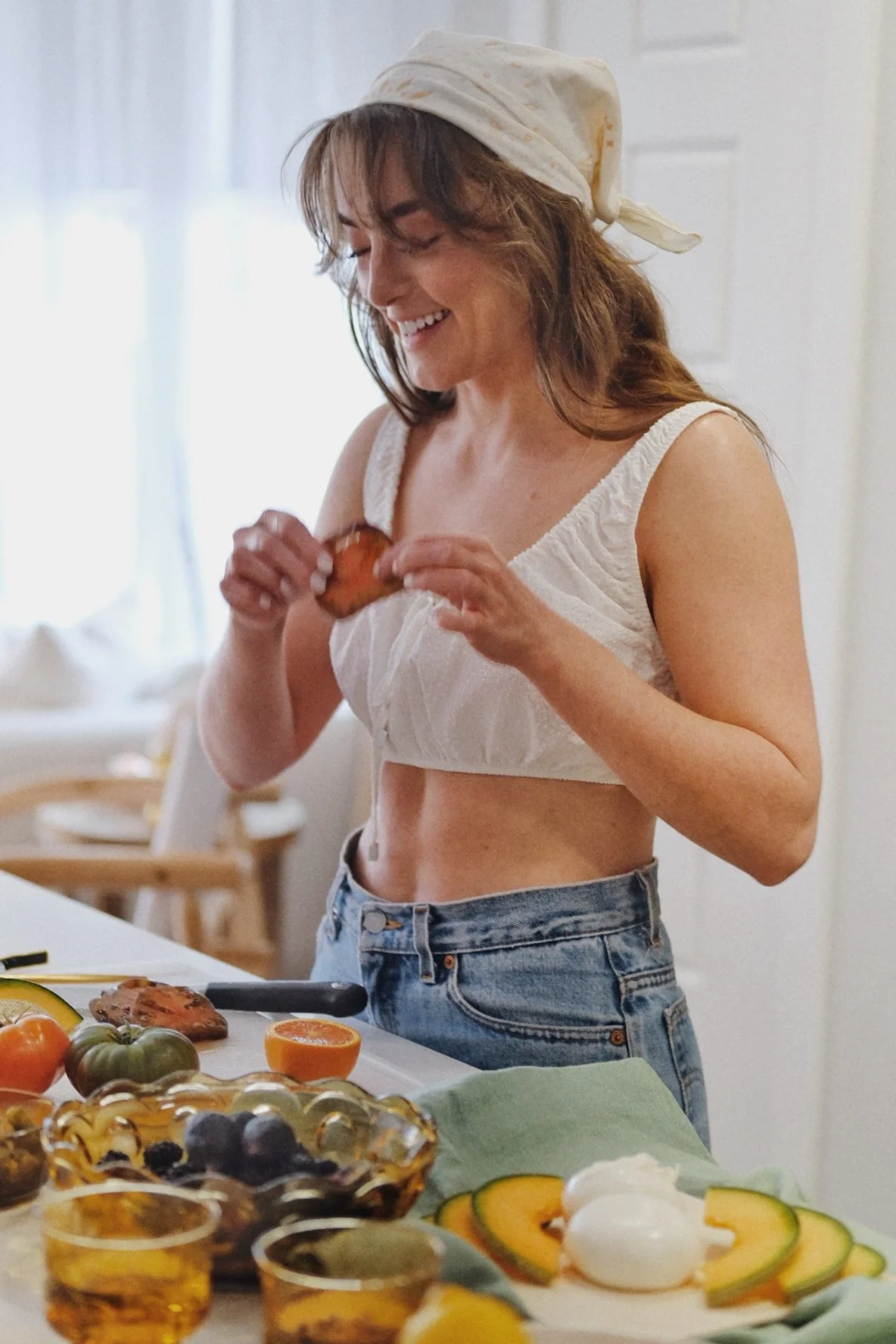 A woman with brown hair wearing a white bandana, white crop top, and jeans, smiling as she holds a piece of food in her hands in a bright kitchen.
