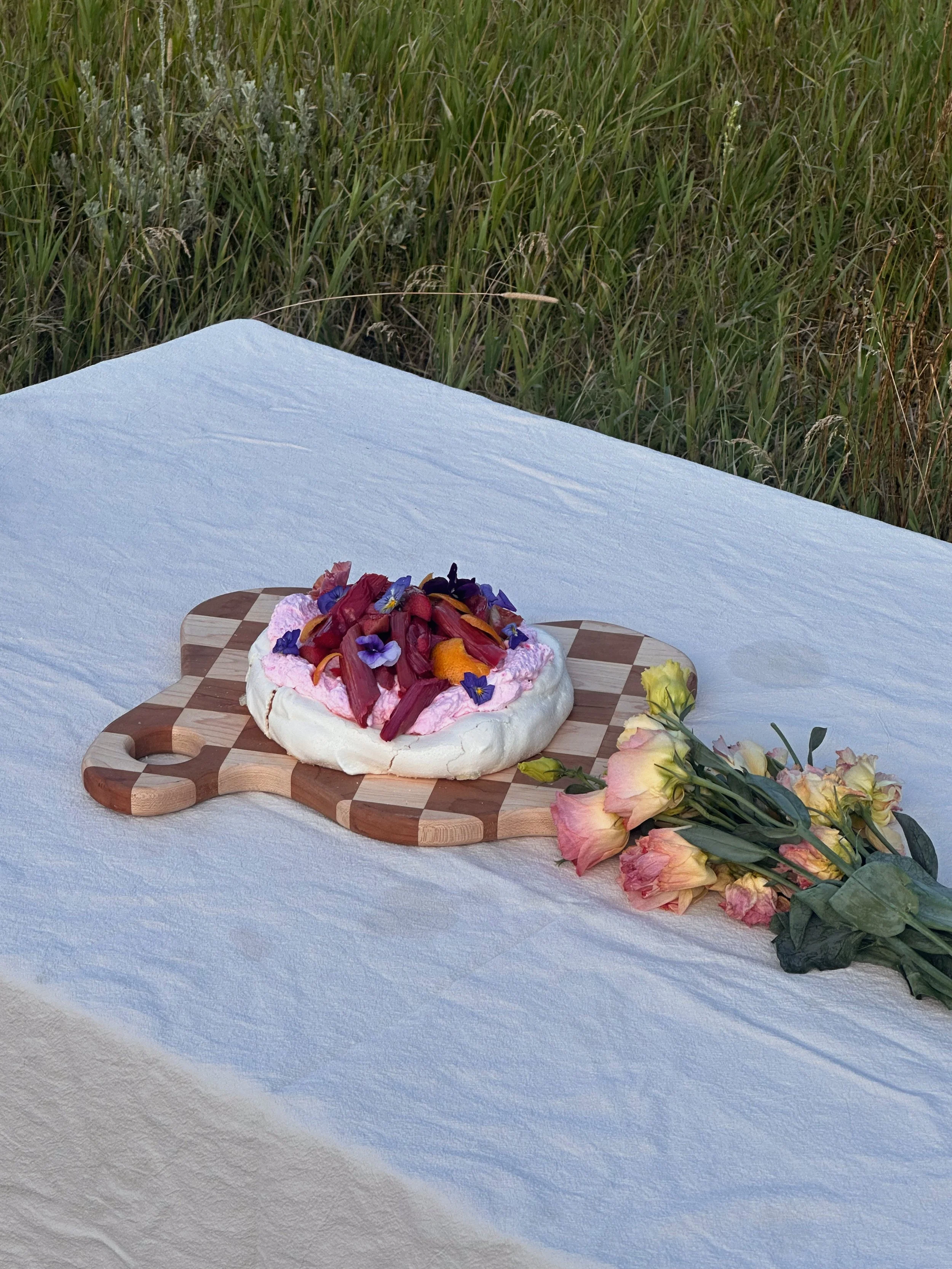 Meringue-based dessert topped with flower petals on a checkered wooden serving board, with a bouquet of pink and yellow roses beside it, set on a white tablecloth outdoors with grass in the background.