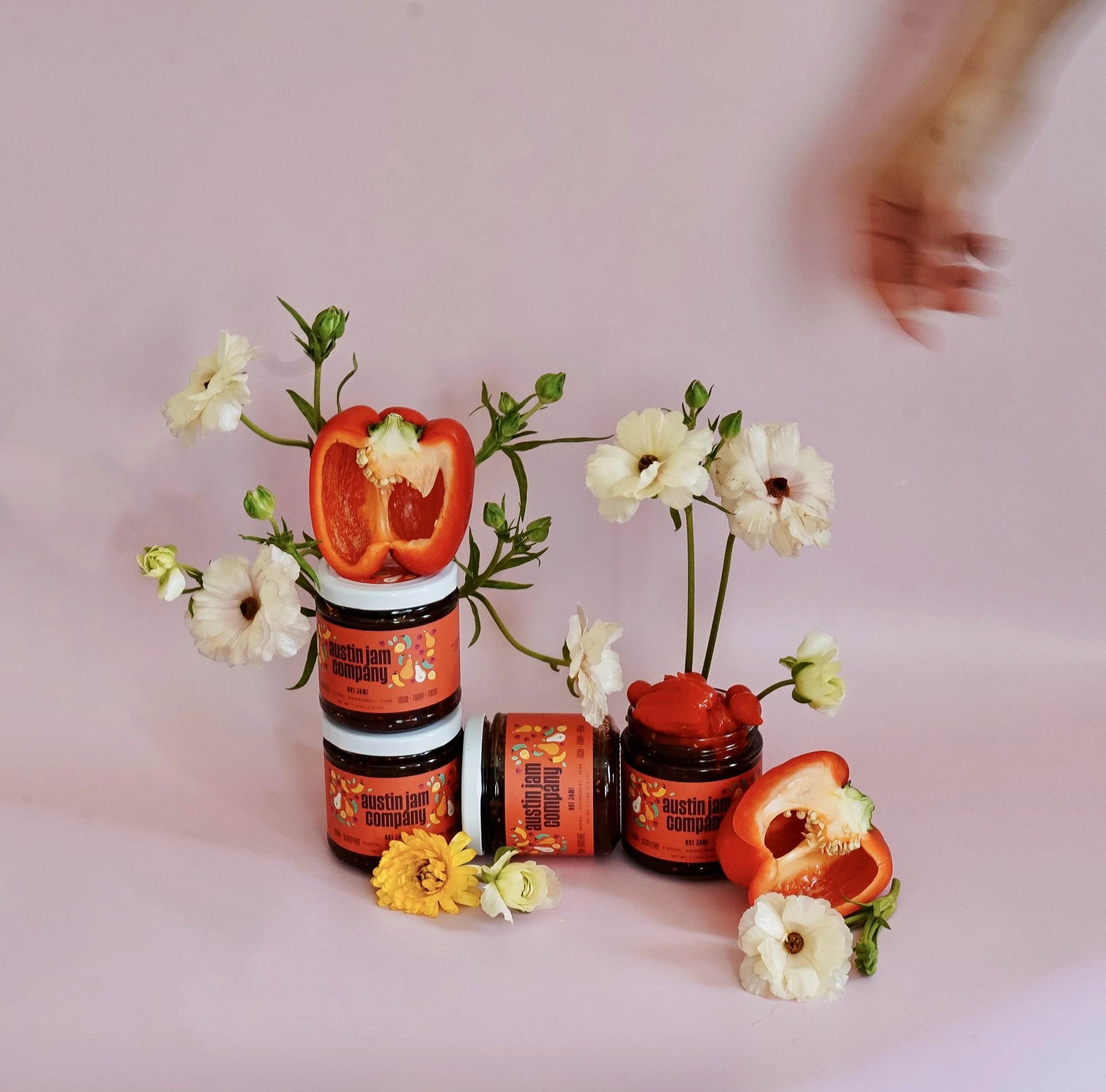 Three jars of Austin Jam Company jam with open red bell peppers on top, surrounded by white and yellow flowers against a pink background, with a blurred hand reaching in from the top right.