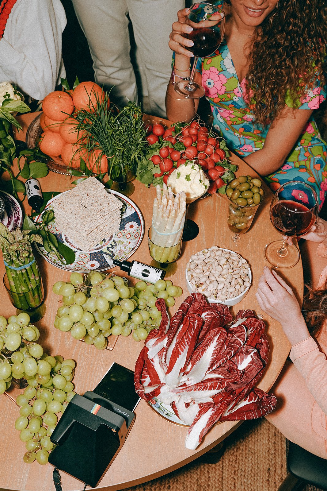 A table with grapes, radishes, olives, pistachios, sliced bread, and various vegetables, with people holding glasses of red wine.