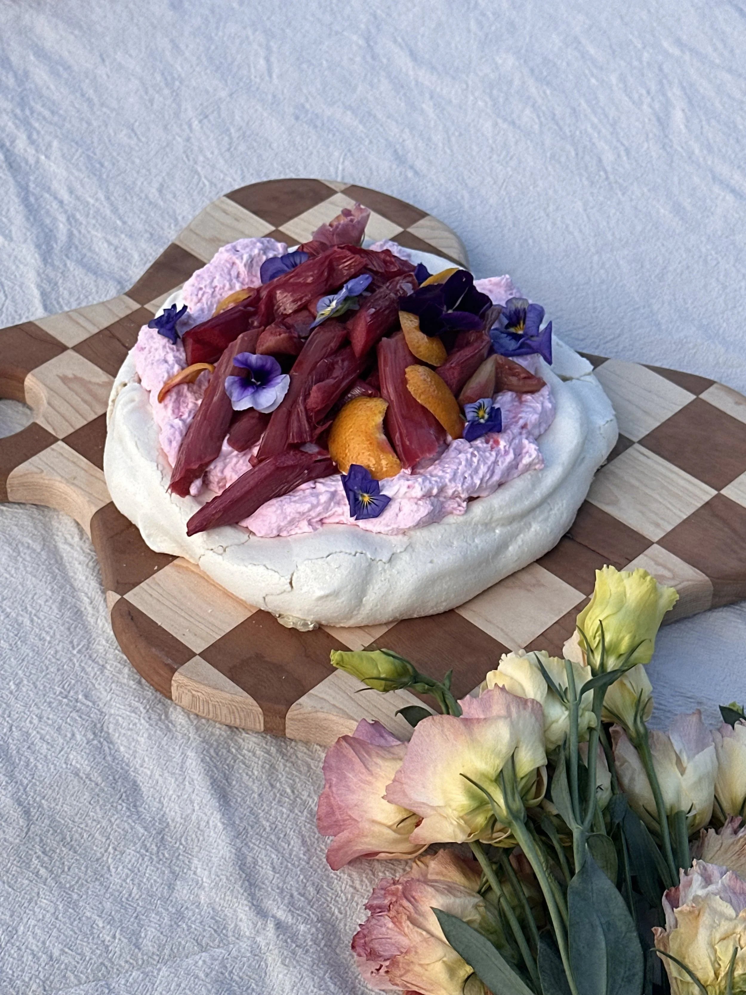 A pavlova topped with whipped cream, pink fruit, orange slices, and edible flowers on a checkered wooden serving board, with a bouquet of pale yellow and pink flowers nearby.