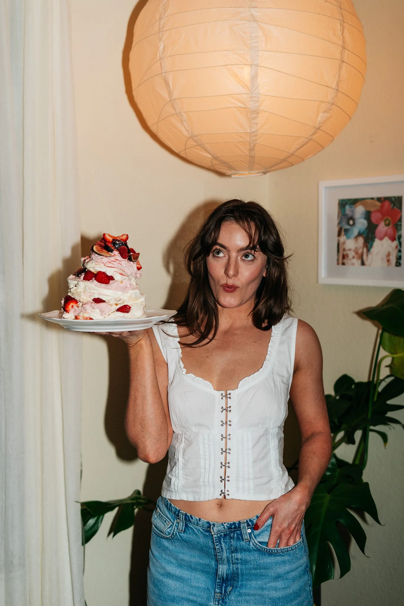 A woman holding a plate with a large dessert topped with strawberries and whipped cream, standing indoors with a large paper lantern overhead and framed floral pictures on the wall.