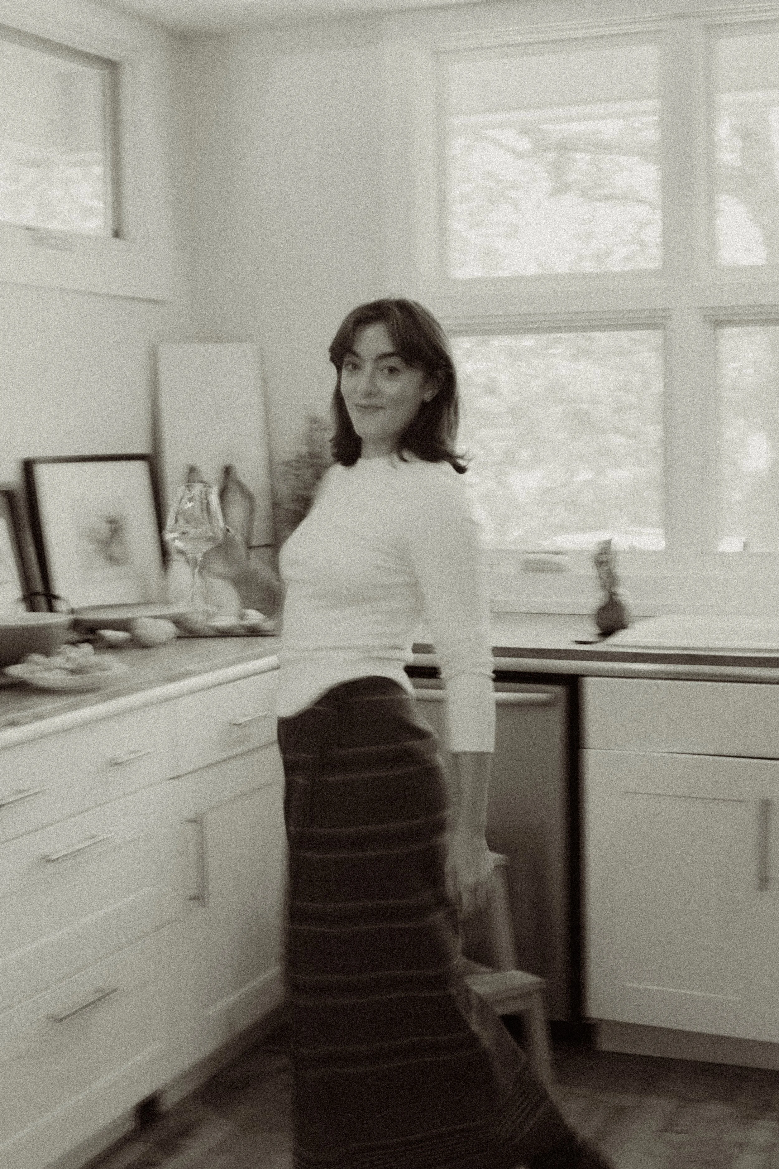 Colette Wetzel standing in a kitchen holding a wine glass, wearing a white top and a striped vintage wool Ralph Lauren skirt, with large windows in the background.