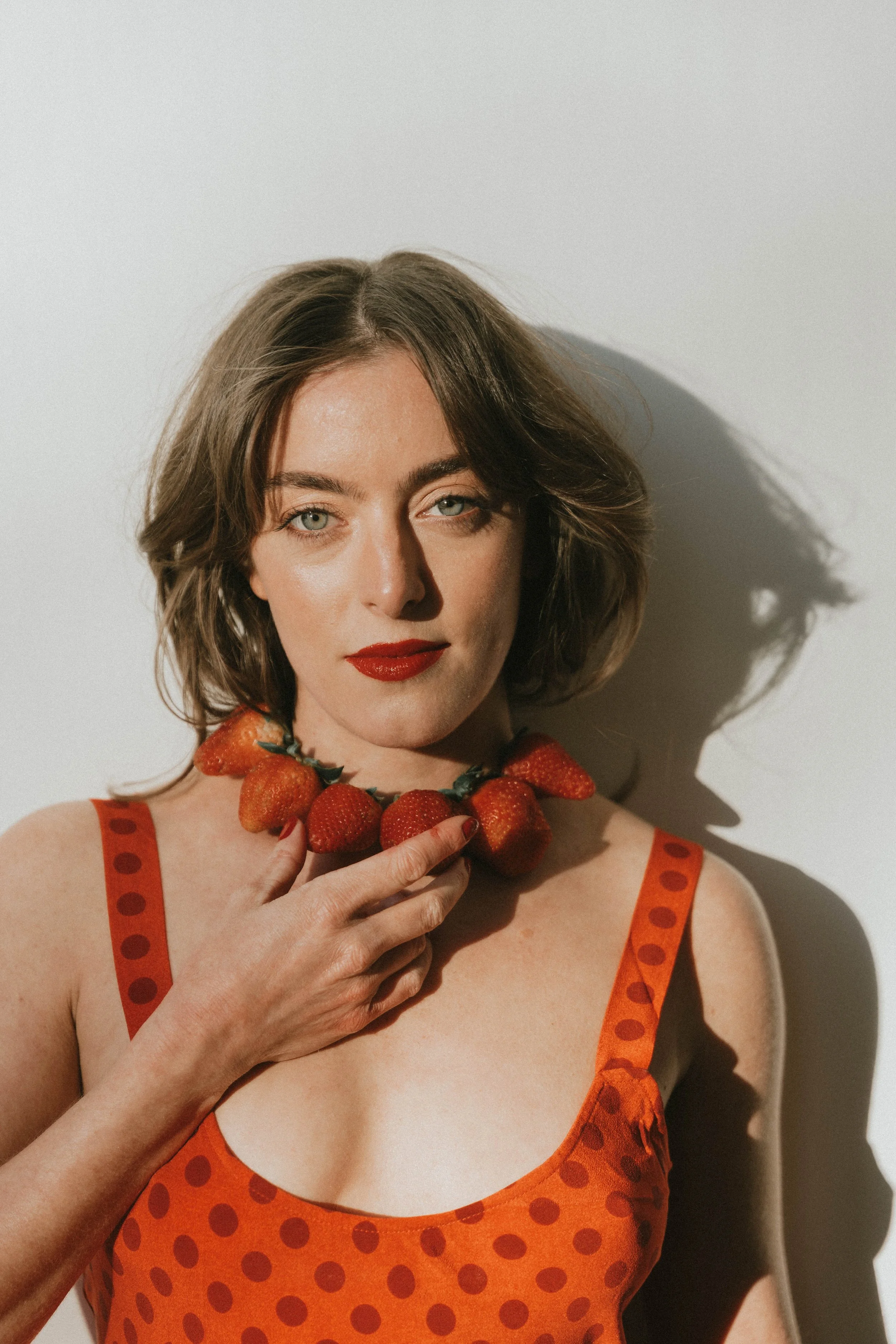 A woman with short brown hair and red lipstick poses against a white background, wearing a red polka-dot dress and a necklace made of strawberries.