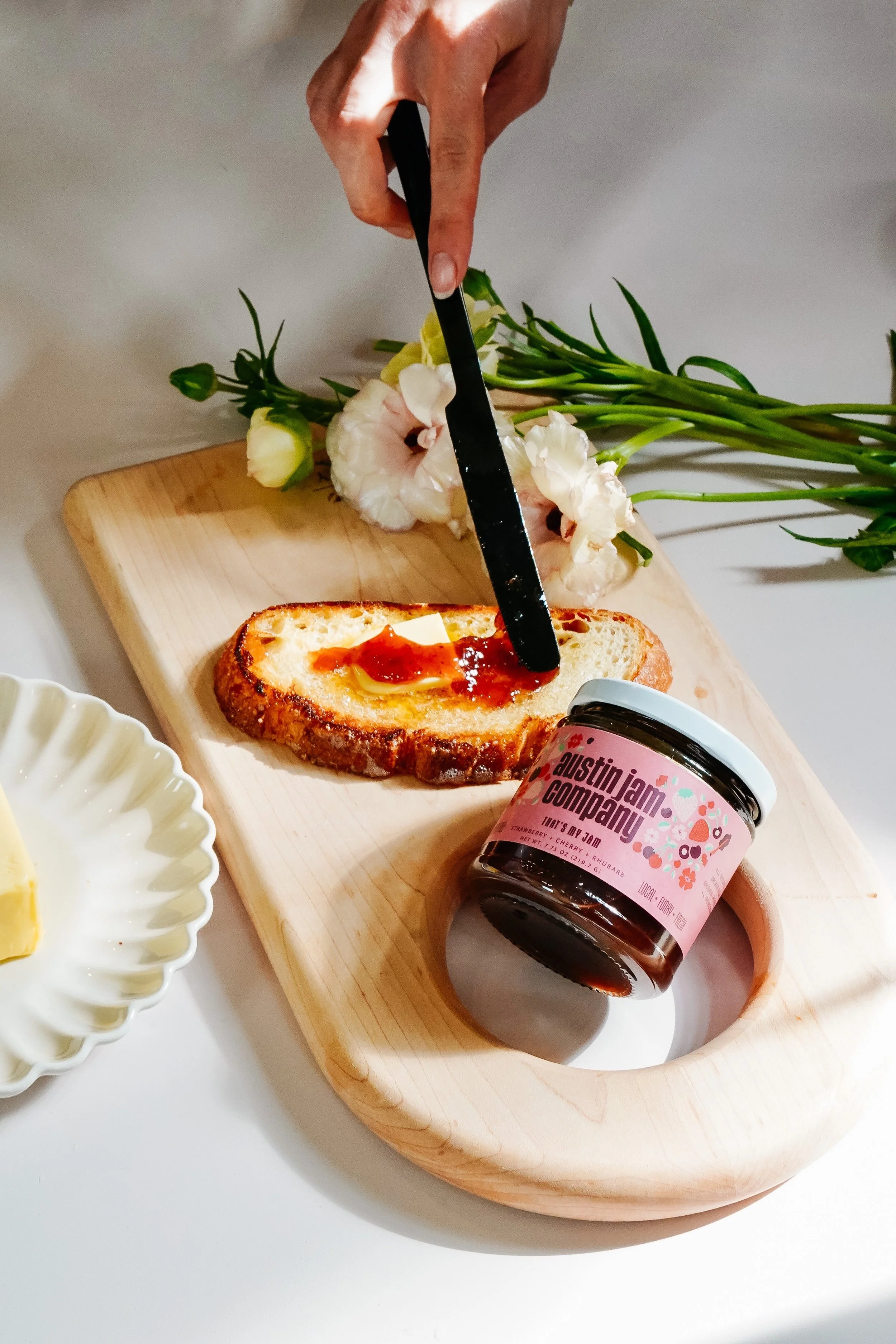A hand spreading jam on a piece of toasted bread with butter, on a wooden serving board that also holds a jar of Austin Jam Company jam and fresh flowers.