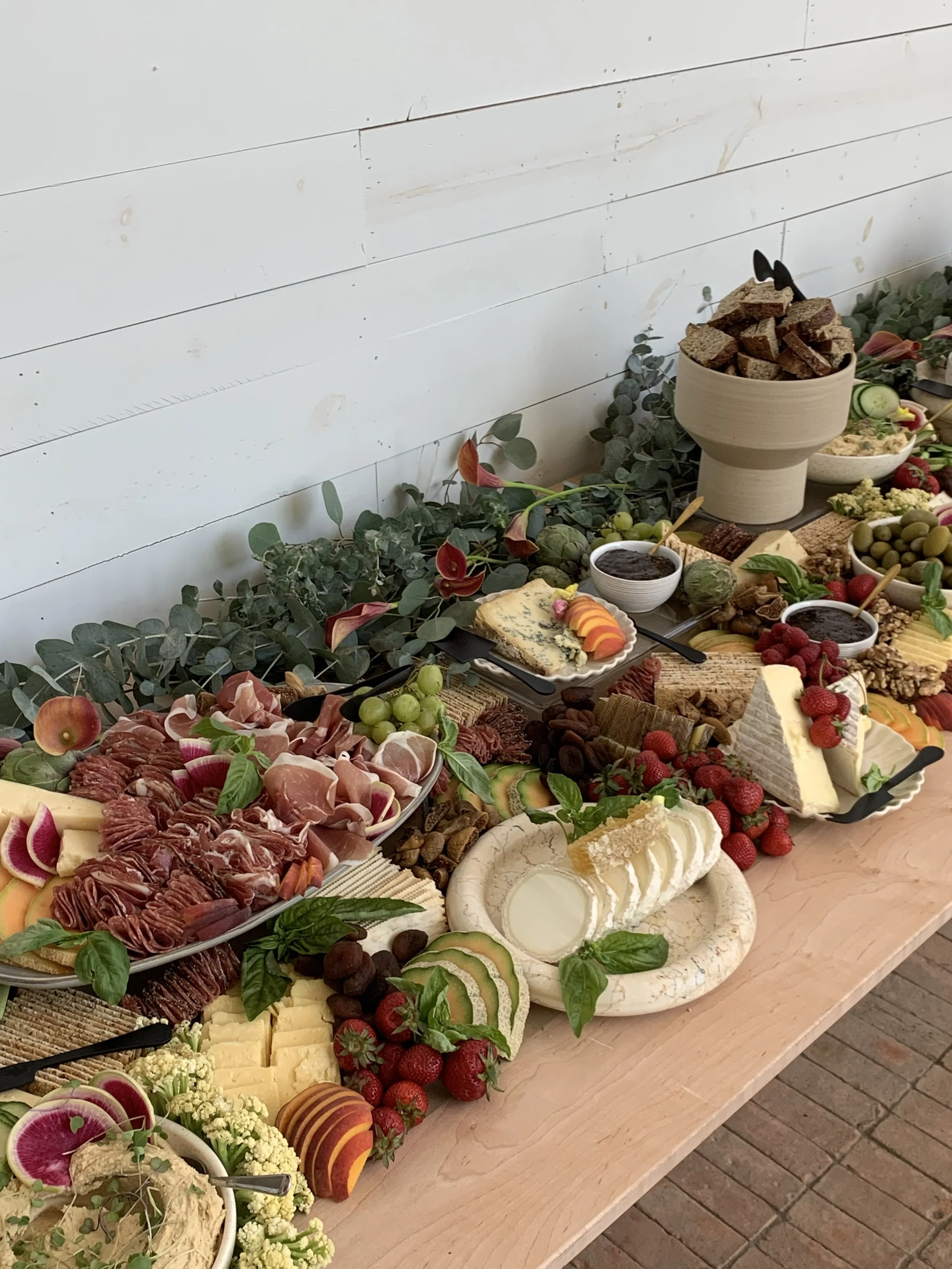 A long table filled with a variety of cheese, charcuterie, crackers, fruits, and dips, decorated with greenery and flowers, set against a white wooden wall.