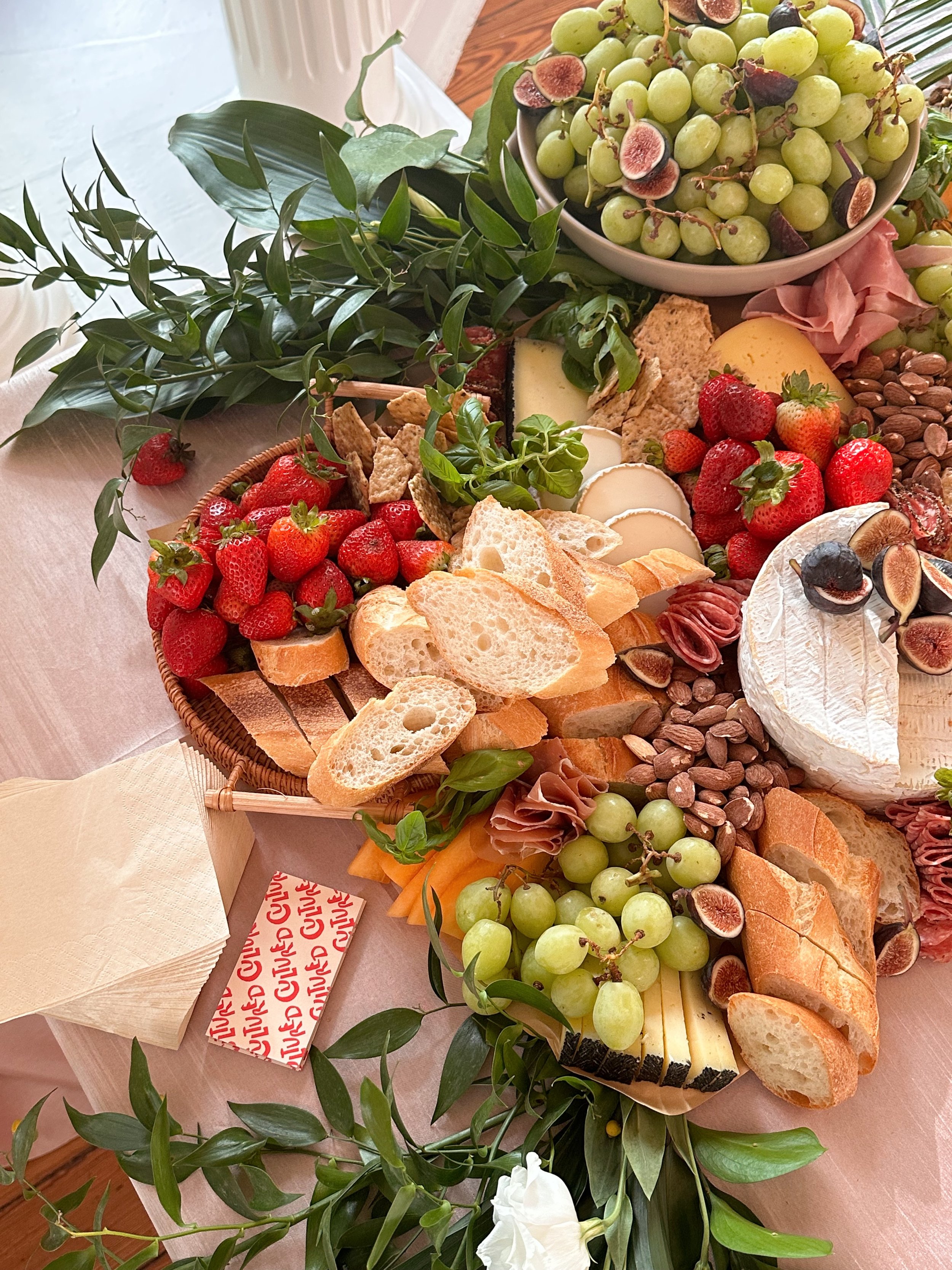 A cheese and fruit platter with strawberries, green grapes, figs, brie cheese, sliced baguette, cured meat, and other cheeses, decorated with green leaves.