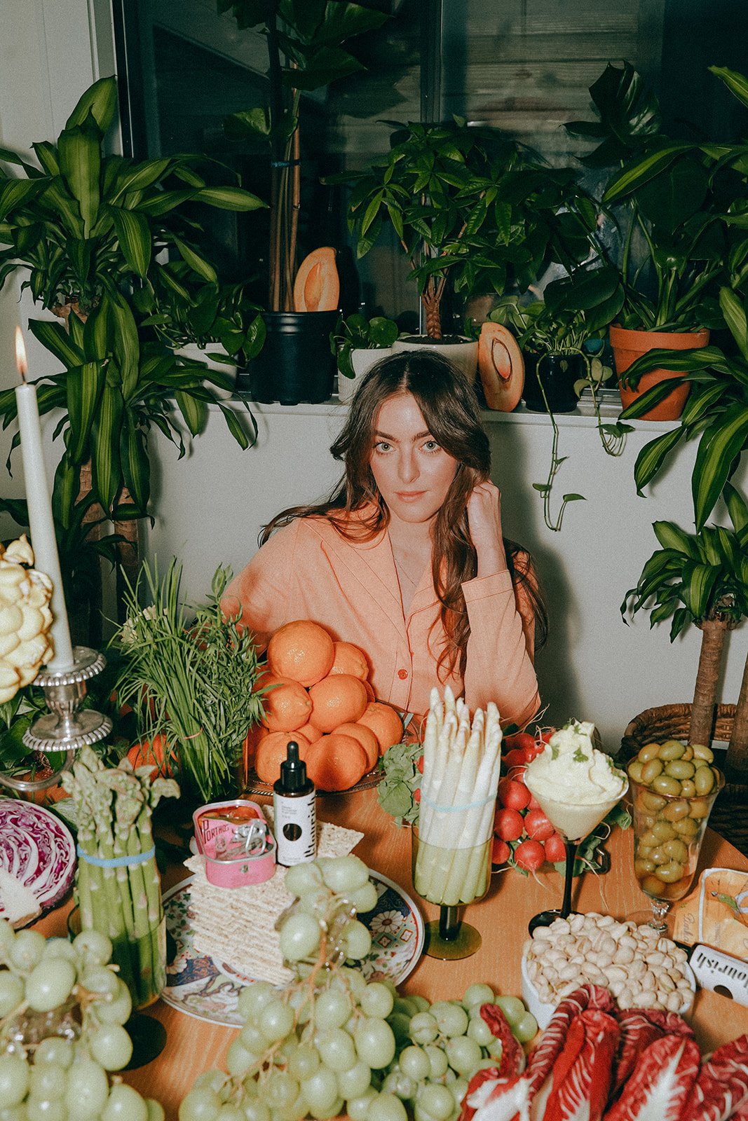 A woman sitting at a dining table surrounded by various fruits and vegetables, including oranges, grapes, tomatoes, and asparagus, with lush green plants in the background.