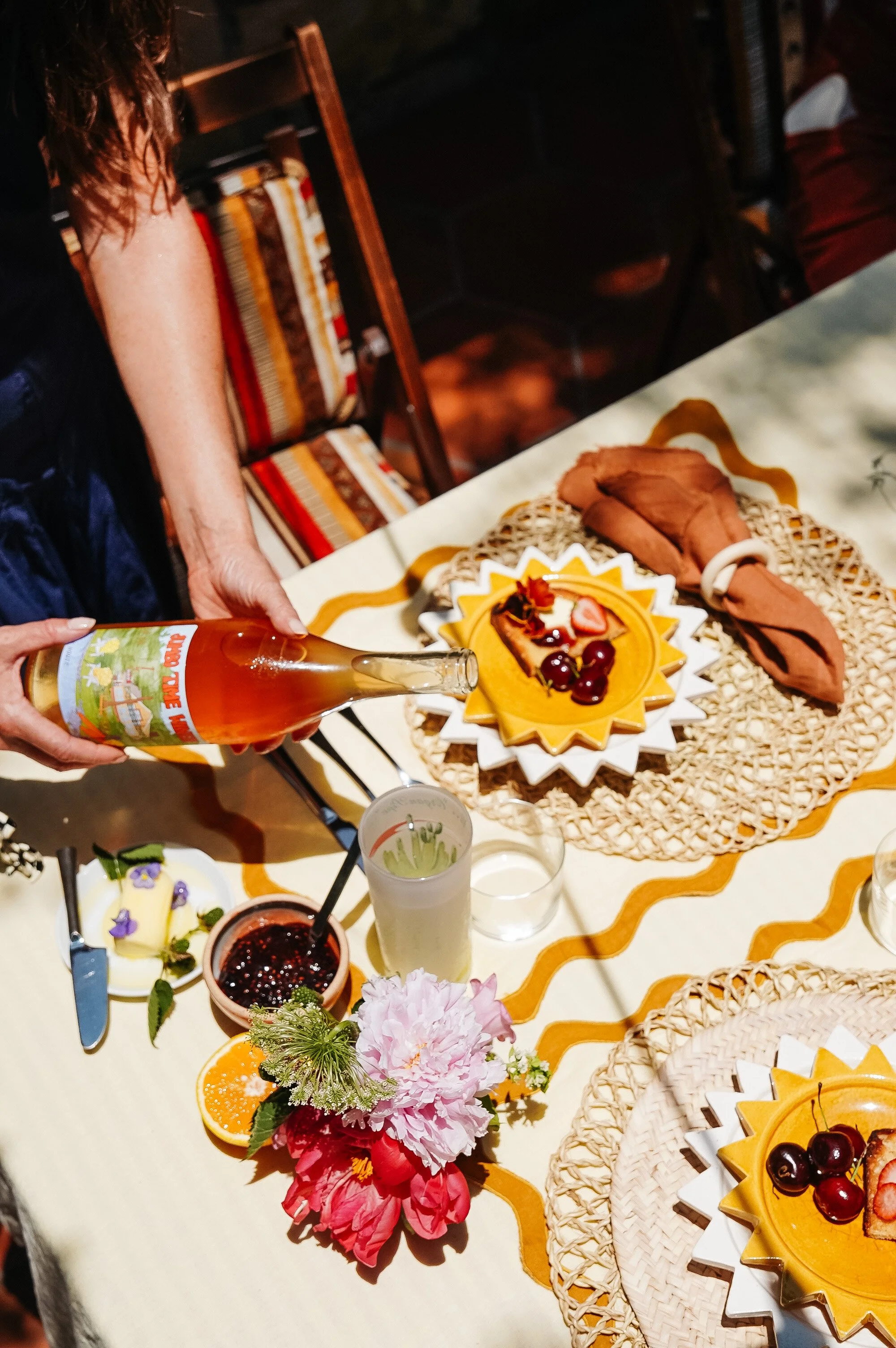 A table with plates of waffles topped with strawberries and cherries, a bottle of rosé wine, a glass of juice, a small vase with pink and red flowers, and various cutlery and fruit slices