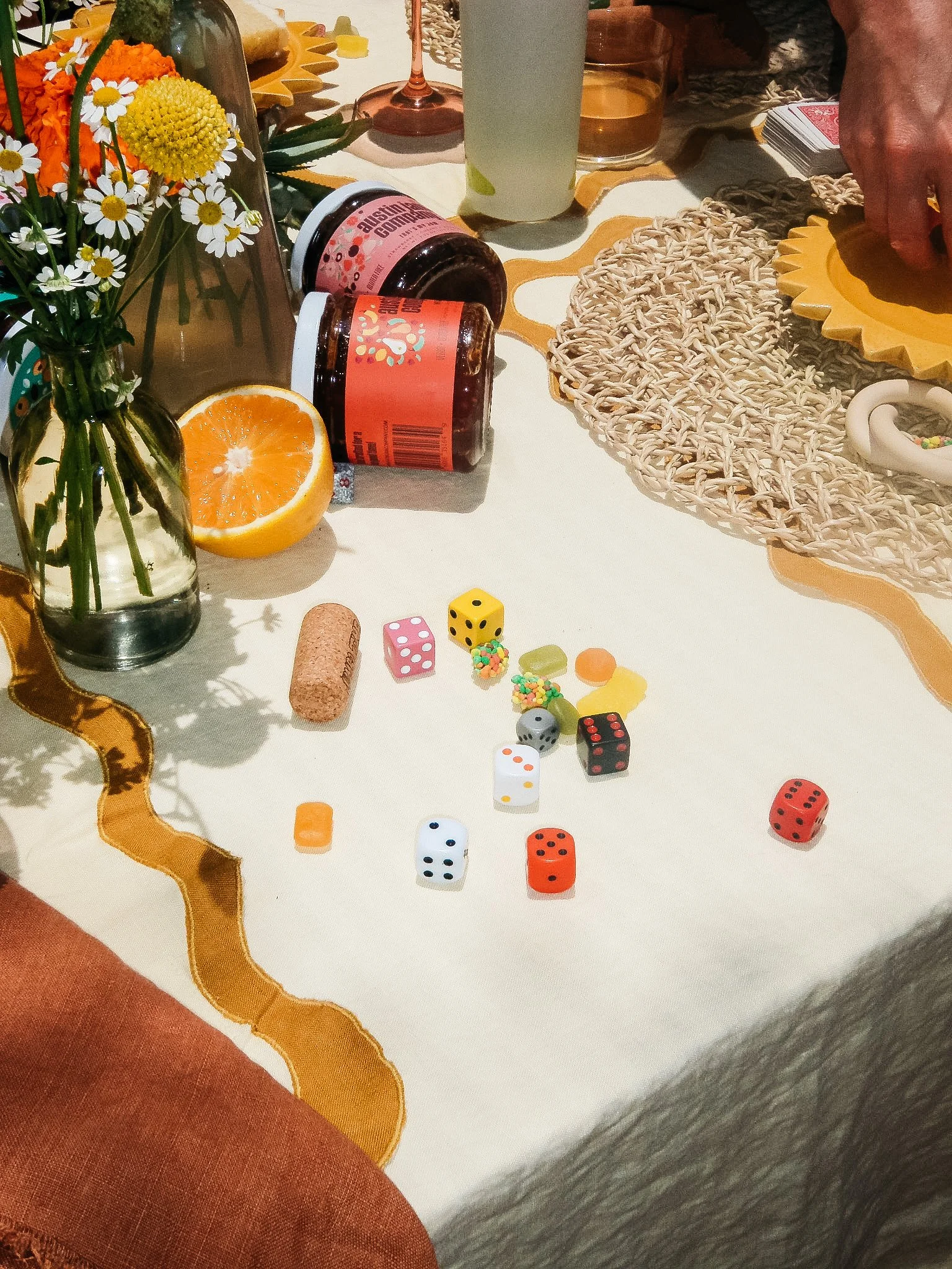 A table with scattered game pieces, including dice and small candies, a half orange, a small bouquet of daisies and orange flowers in a glass vase, bottles of medication, and various glasses with drinks.
