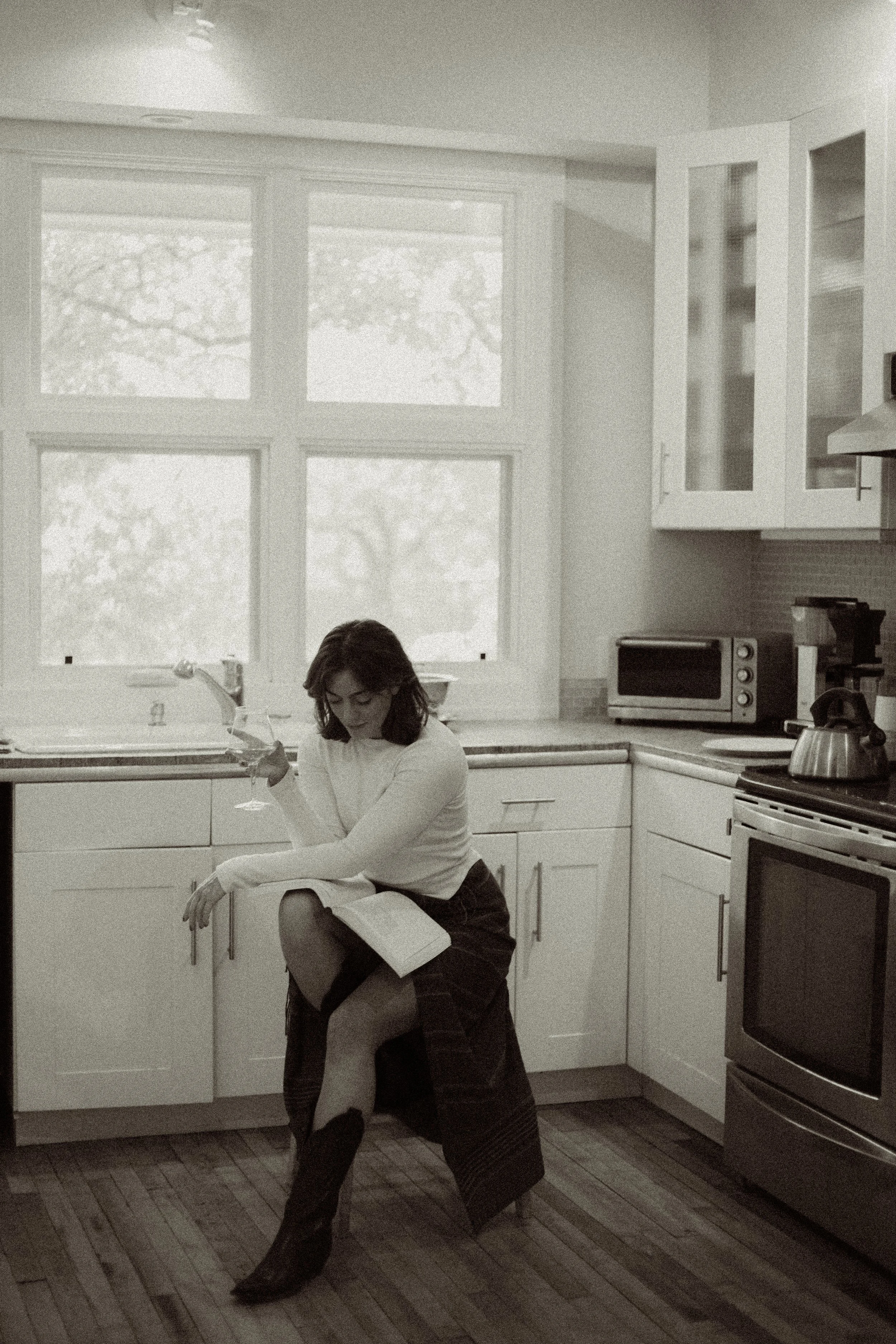 A woman sitting on a kitchen counter in a modern kitchen, looking down at a drink in her hand. She is wearing a white top, a plaid skirt, and cowboy boots, with her other leg resting on the counter.