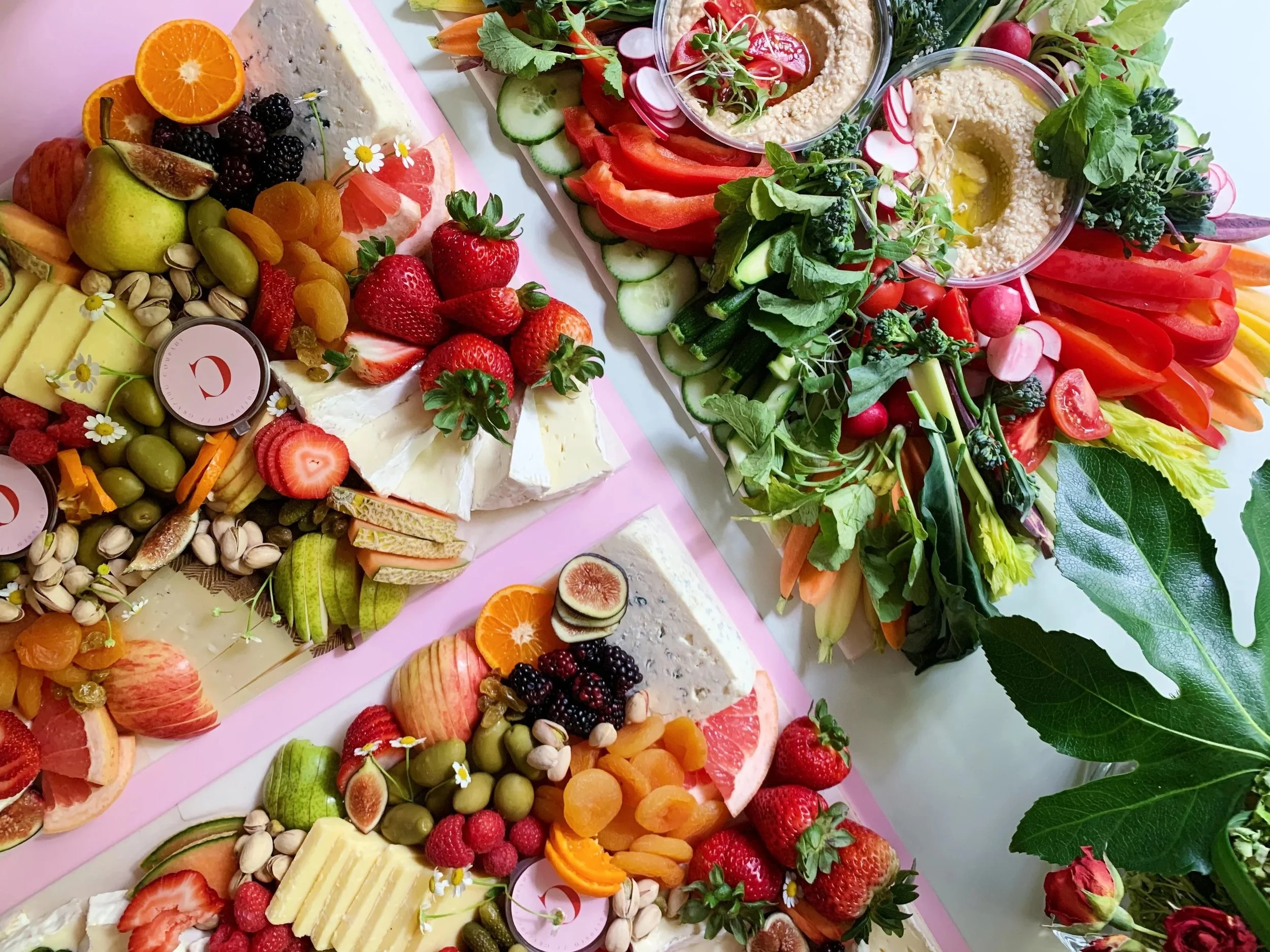 Cultured Grazing Company. Colorful arrangement of cheese, fresh fruits, vegetables, and hummus with vegetables garnished with radishes, cherry tomatoes, and leafy greens.