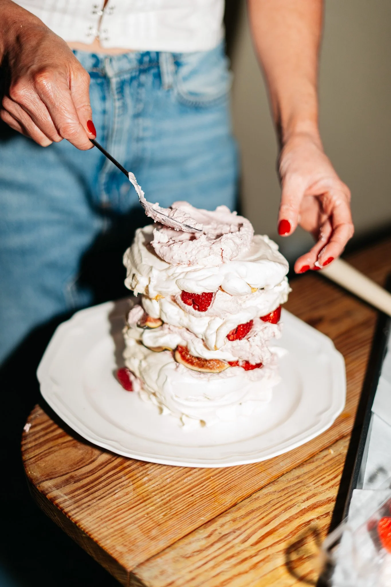 A person wearing a white top and blue jeans is assembling a layered dessert with whipped cream, strawberries, figs, and meringue on a white plate on a wooden table.