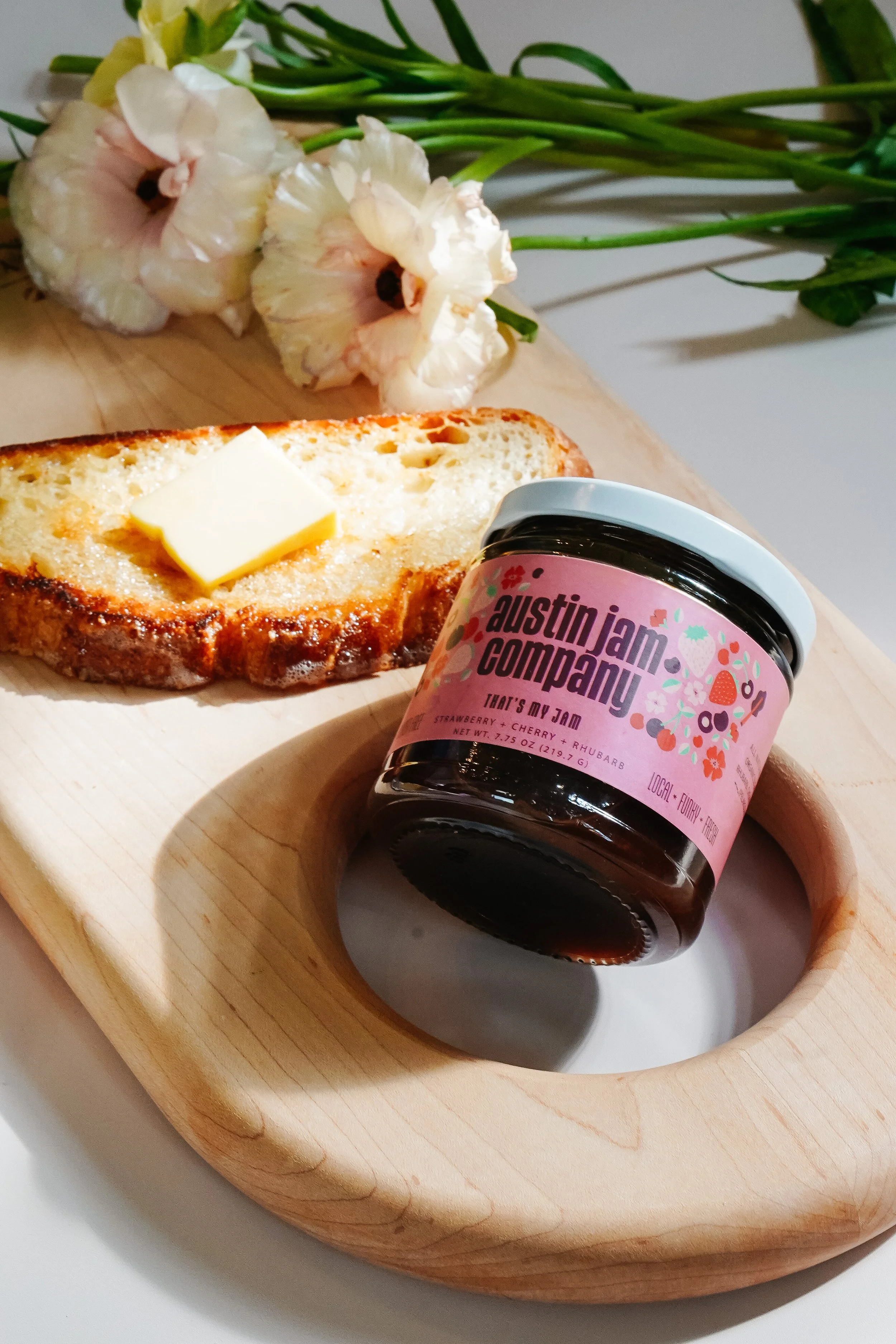 A wooden tray holding a slice of bread with butter, a jar of jam labeled 'Austin Jam Company' with strawberry, cherry, and rhubarb flavors, and a bouquet of cream-colored flowers in the background.