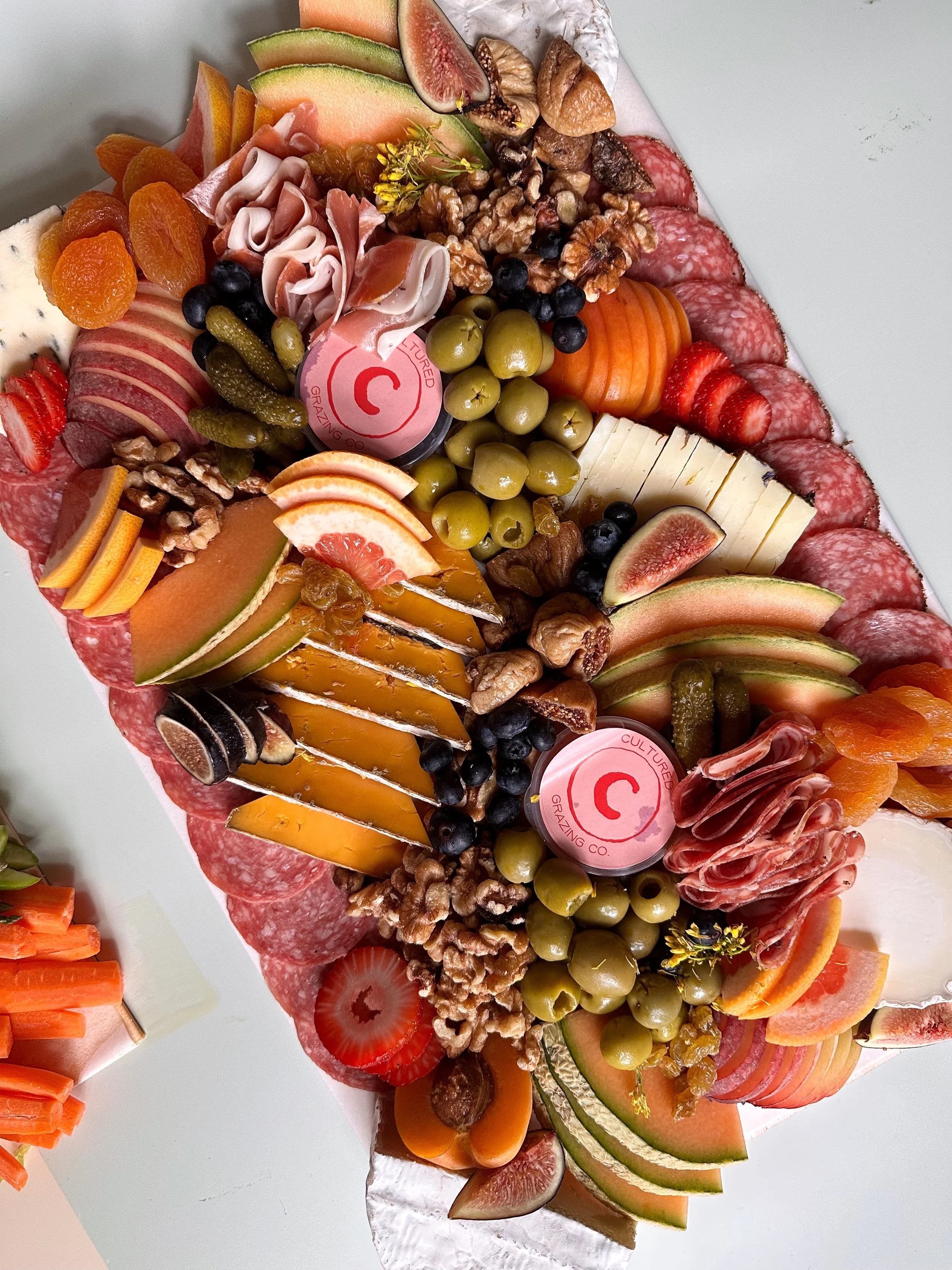 A charcuterie board with assorted cheeses, cured meats, fresh fruits, nuts, and pickles on a white surface.